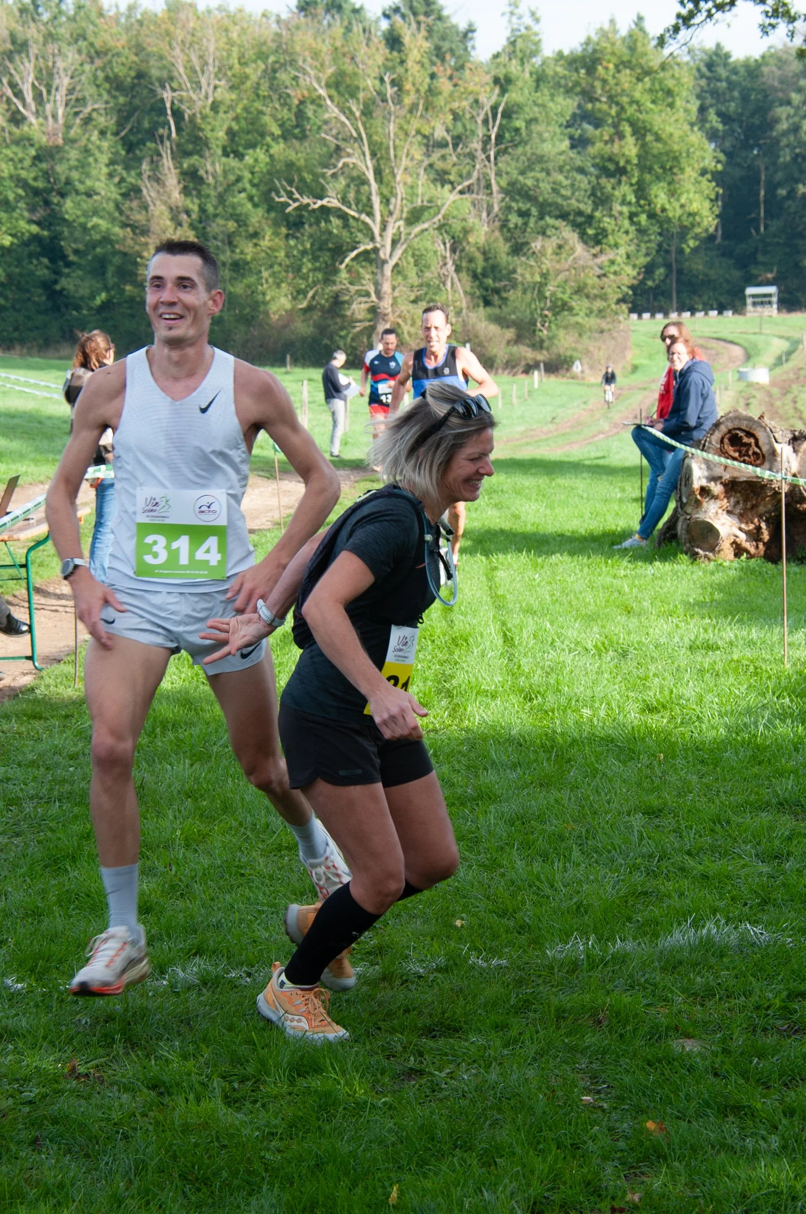 Deux coureurs, un homme en débardeur blanc et une femme en t-shirt noir, célèbrent à la fin d'une course en extérieur dans un parc vert, avec d'autres participants visibles en arrière-plan.