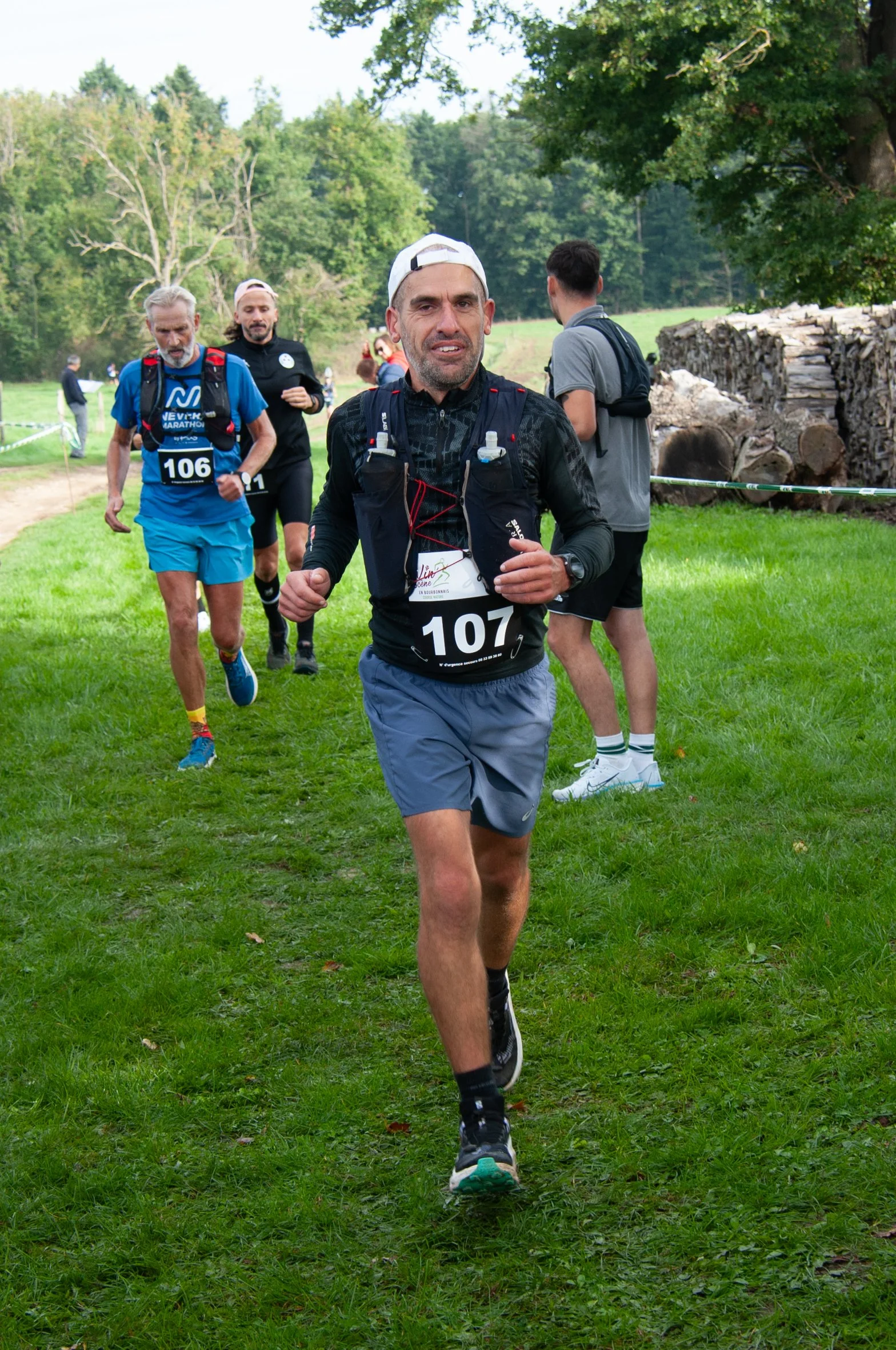 Des coureurs participant à une course en extérieur, portant des dossards avec les numéros 107, 106 et d'autres, sur un parcours herbeux avec des arbres en arrière-plan.
