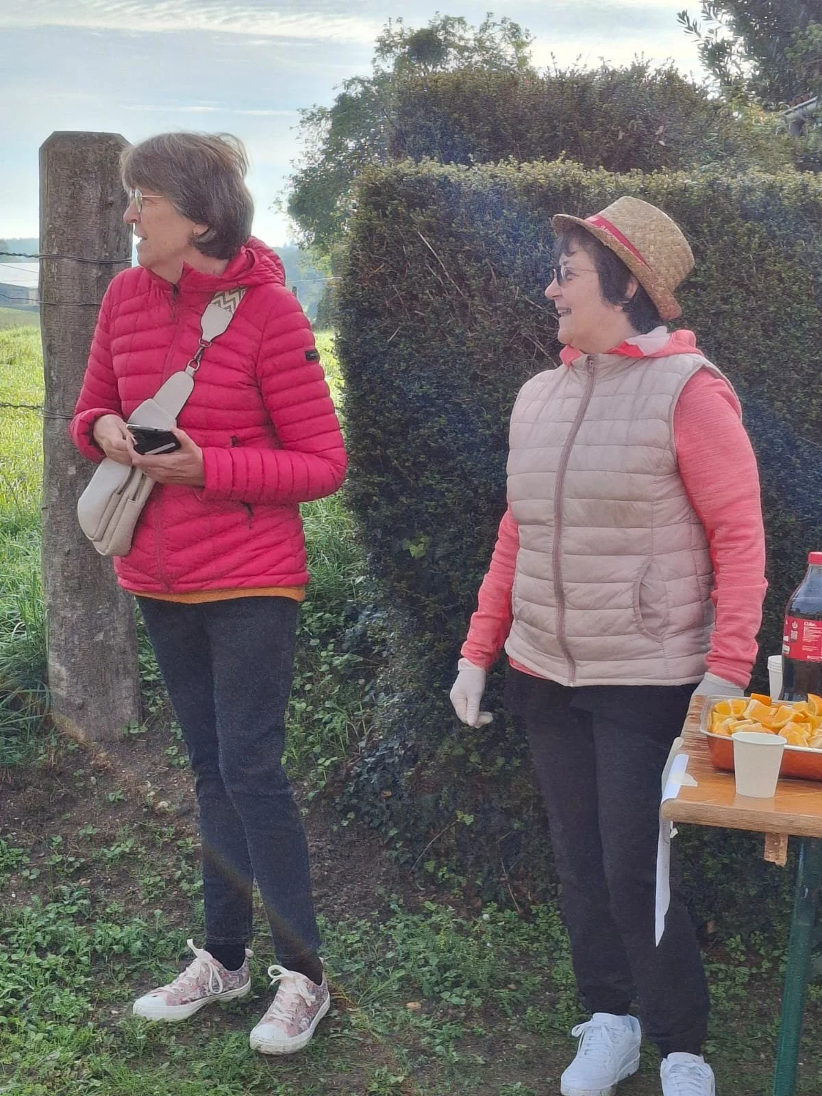 Deux femmes souriantes, portant des vêtements décontractés et des vestes, se tiennent près d'une table avec des boissons et des snacks, dans un environnement extérieur avec des buissons et un ciel partiellement nuageux.