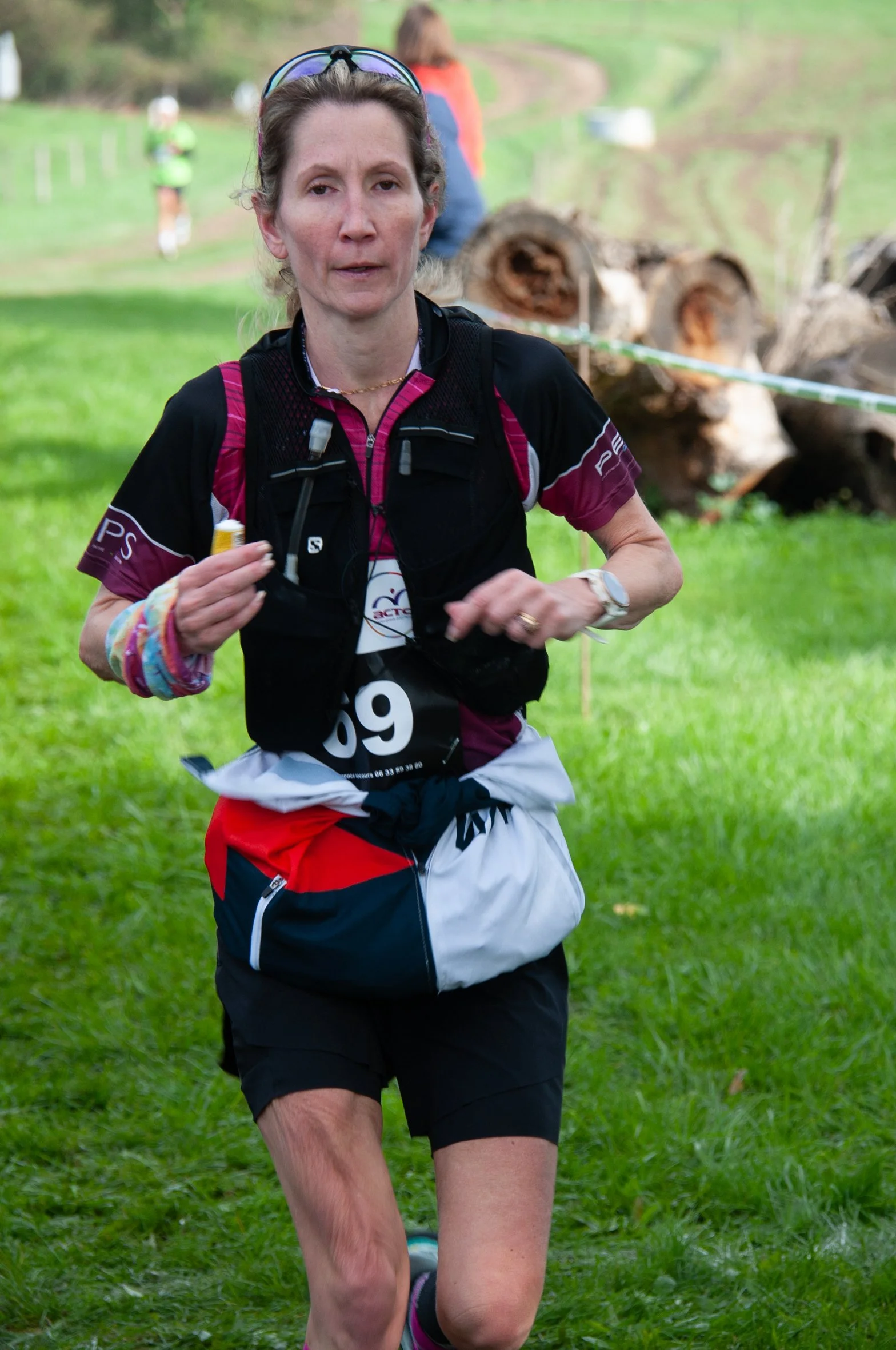 Femme courant lors d'une course en plein air avec un numéro 69, portant un sac à taille, des vêtements de sport colorés, et des lunettes sur la tête, dans un cadre naturel avec de l'herbe verte et des troncs d'arbres en arrière-plan.