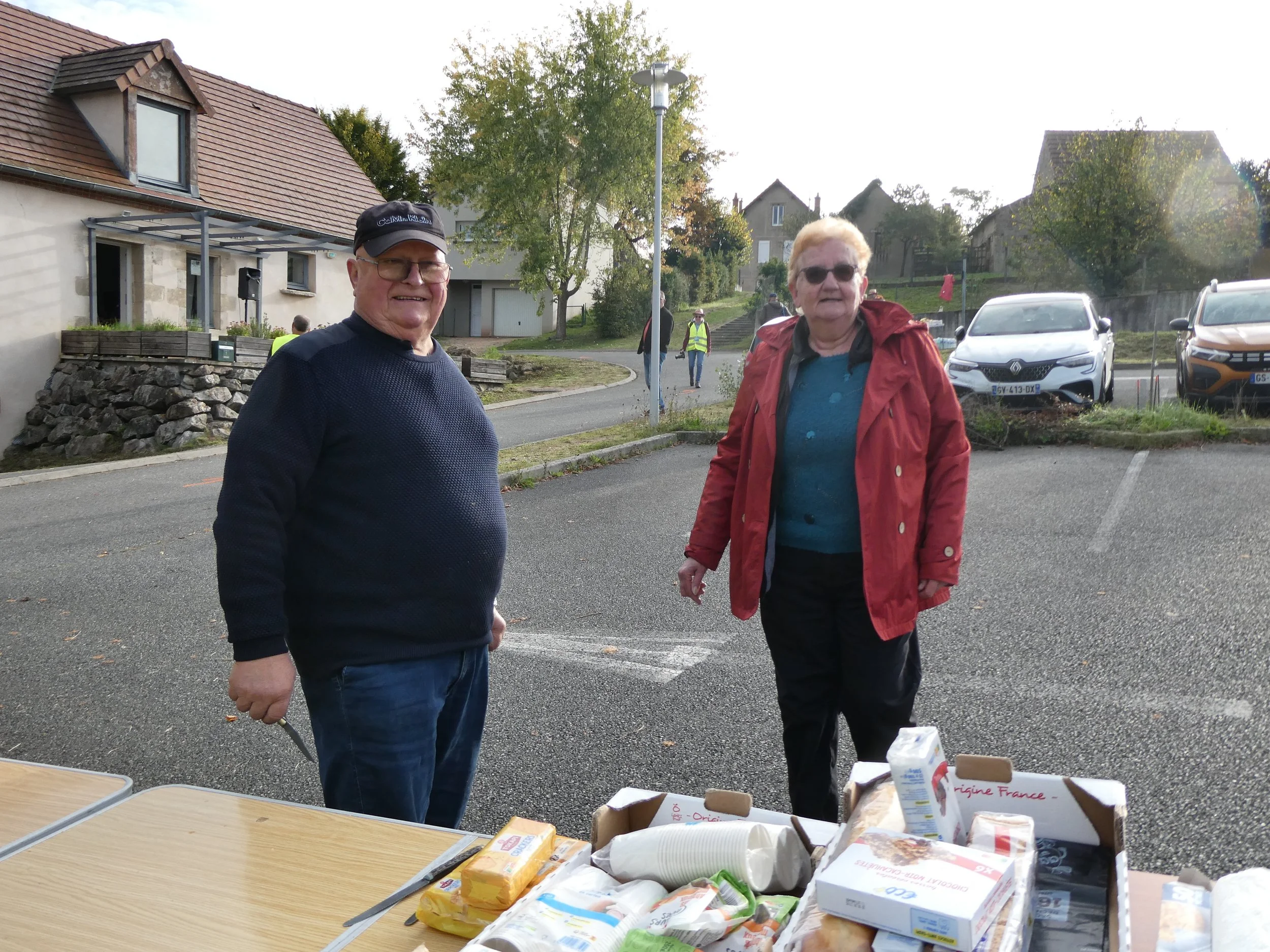 Deux personnes âgées sont debout près d'une table contenant des produits alimentaires, dans une place de stationnement en plein air, avec des maisons en arrière-plan, en plein jour.