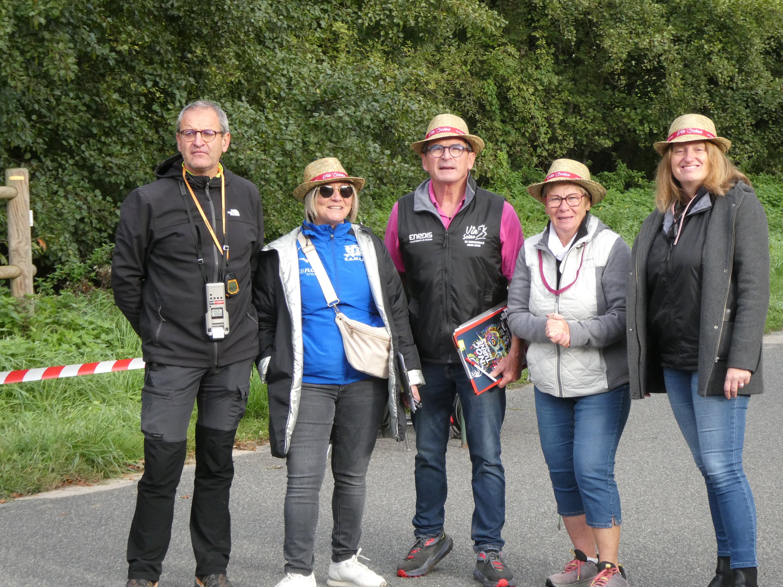 Groupe de cinq personnes posant pour une photo en plein air, certaines portant des chapeaux en paille et des vestes, dans un environnement verdoyant.