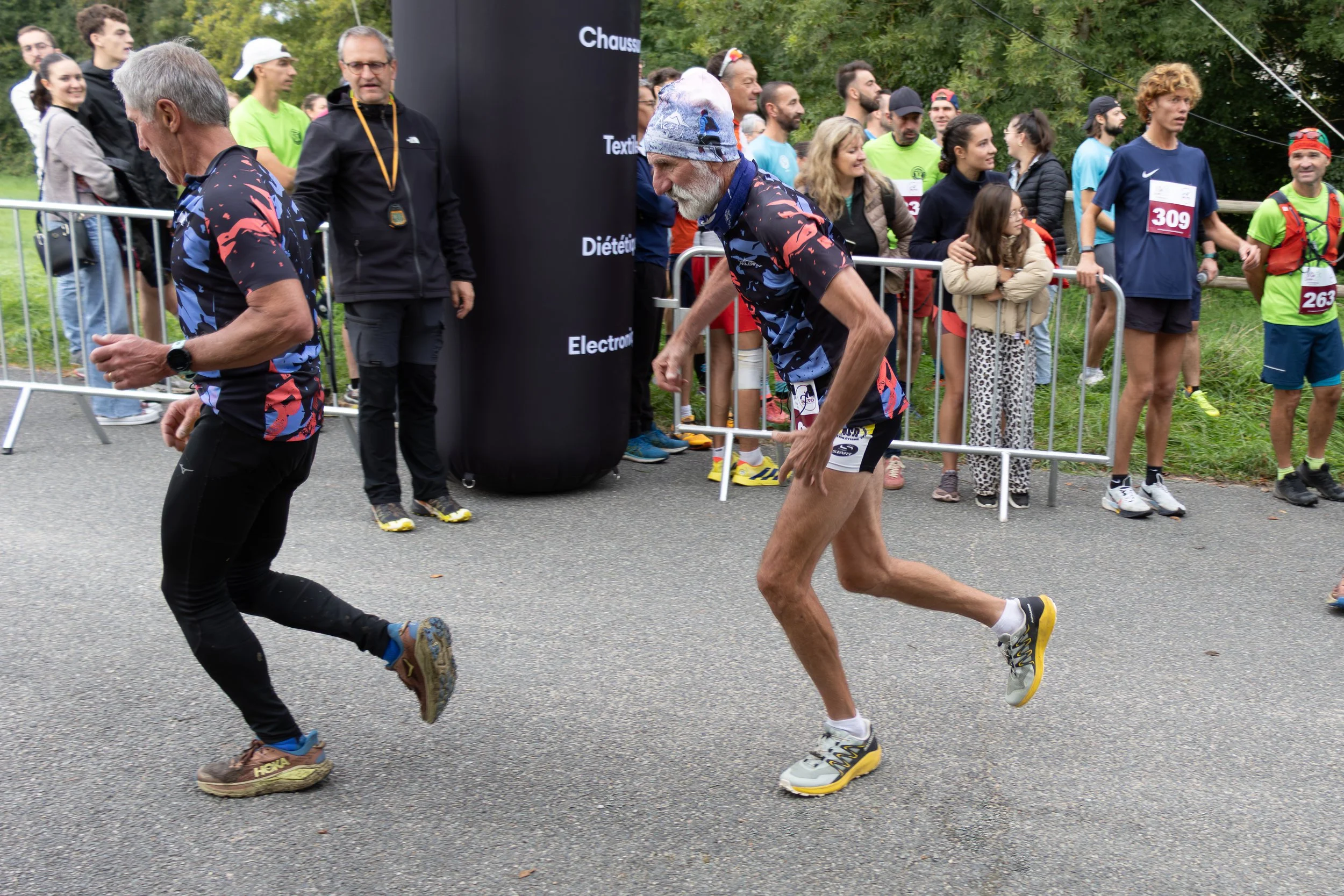 Deux coureurs, dont un vieux homme, en train de courir lors d'une compétition, avec des spectateurs derrière une barrière.