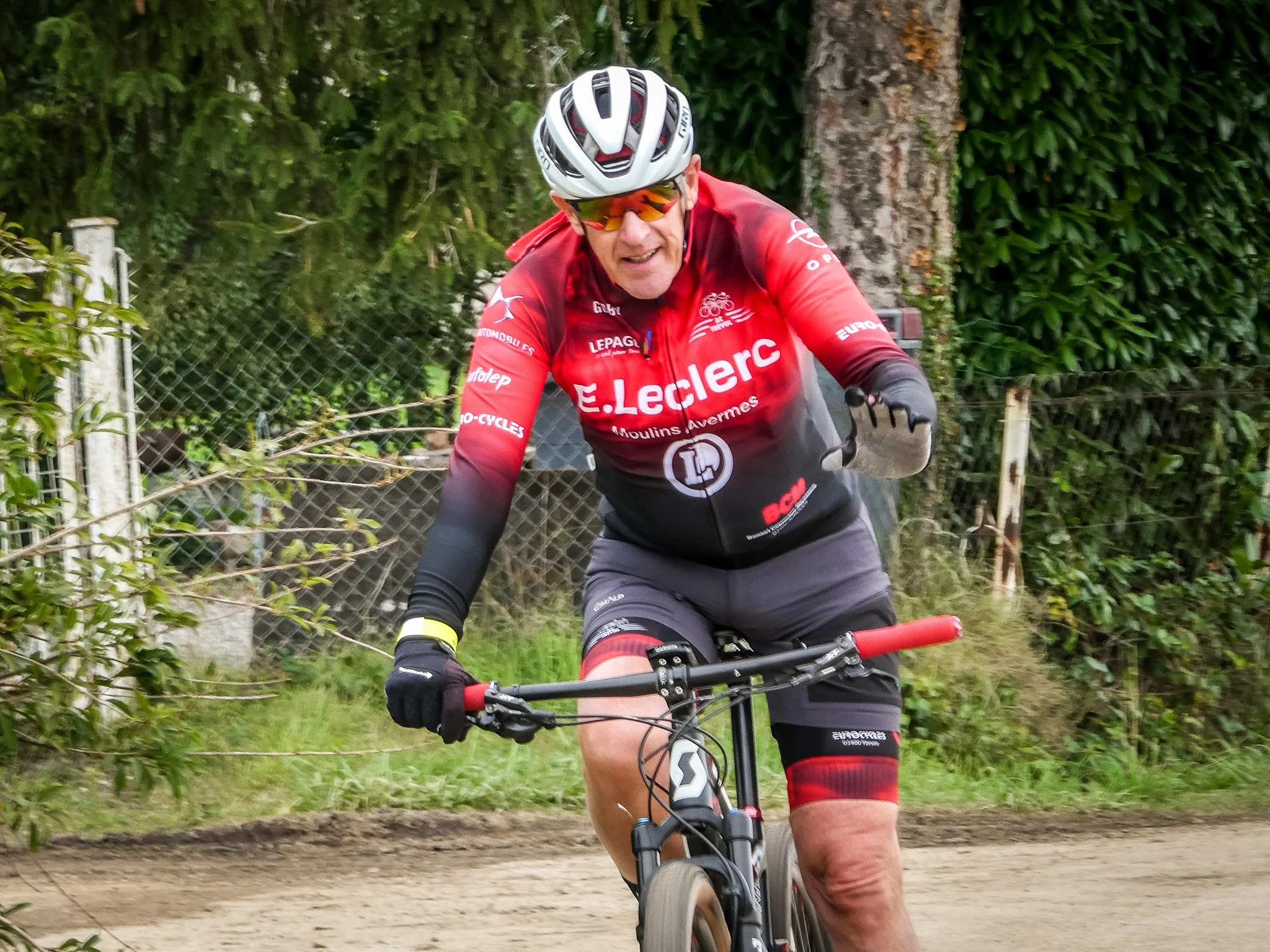 Un homme en maillot de cyclisme rouge, portant un casque et des lunettes, fait du vélo sur un chemin en extérieur entouré de végétation.