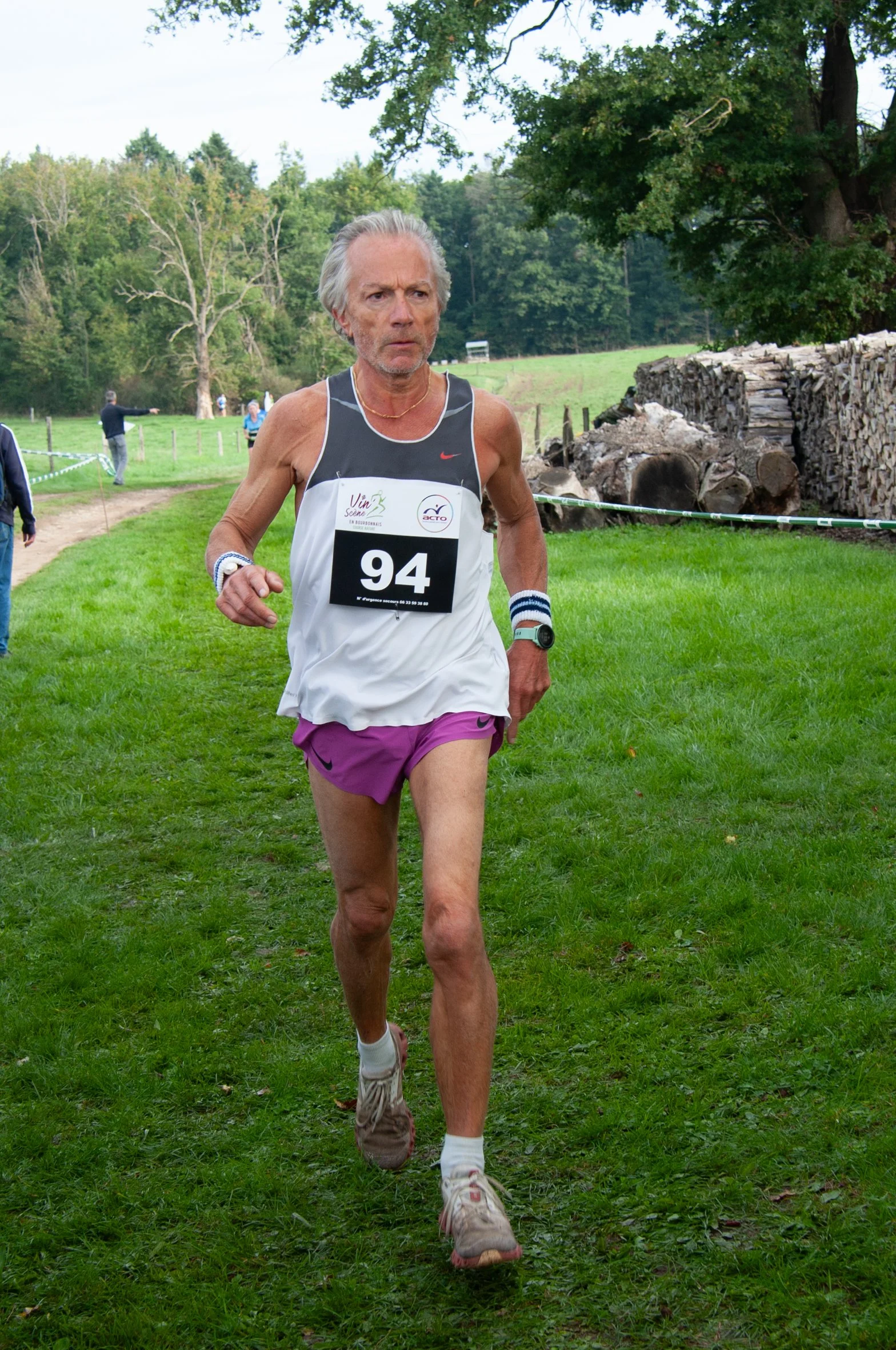 Un homme âgé court lors d'une course en plein air dans un environnement naturel avec des arbres et des bûches de bois en arrière-plan, portant un dossard numéro 94, un maillot blanc, des shorts violets, et des bracelets aux poignets.