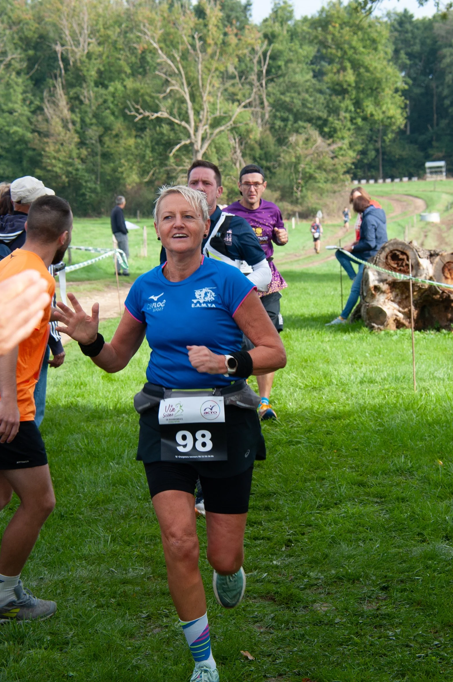 Une femme courrant lors d'une course en plein air, portant un maillot bleu, le numéro 98, avec plusieurs personnes en arrière-plan, sur un terrain verdoyant entouré d'arbres.