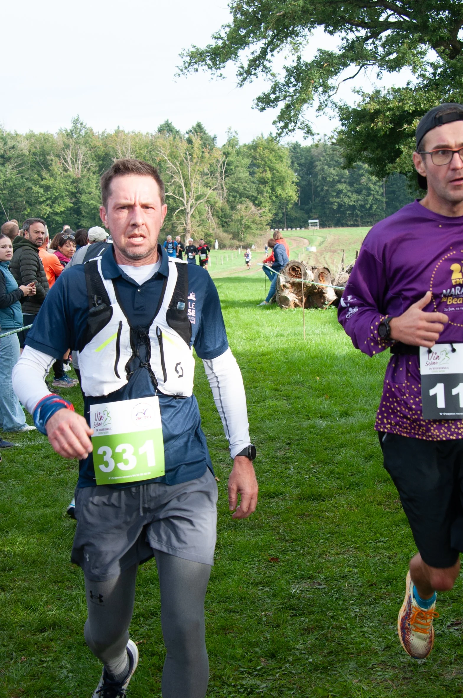Deux hommes courant lors d'une course de trail dans un parc vert, avec d'autres coureurs et spectateurs en arrière-plan, entouré d'arbres et d'une nature luxuriante.