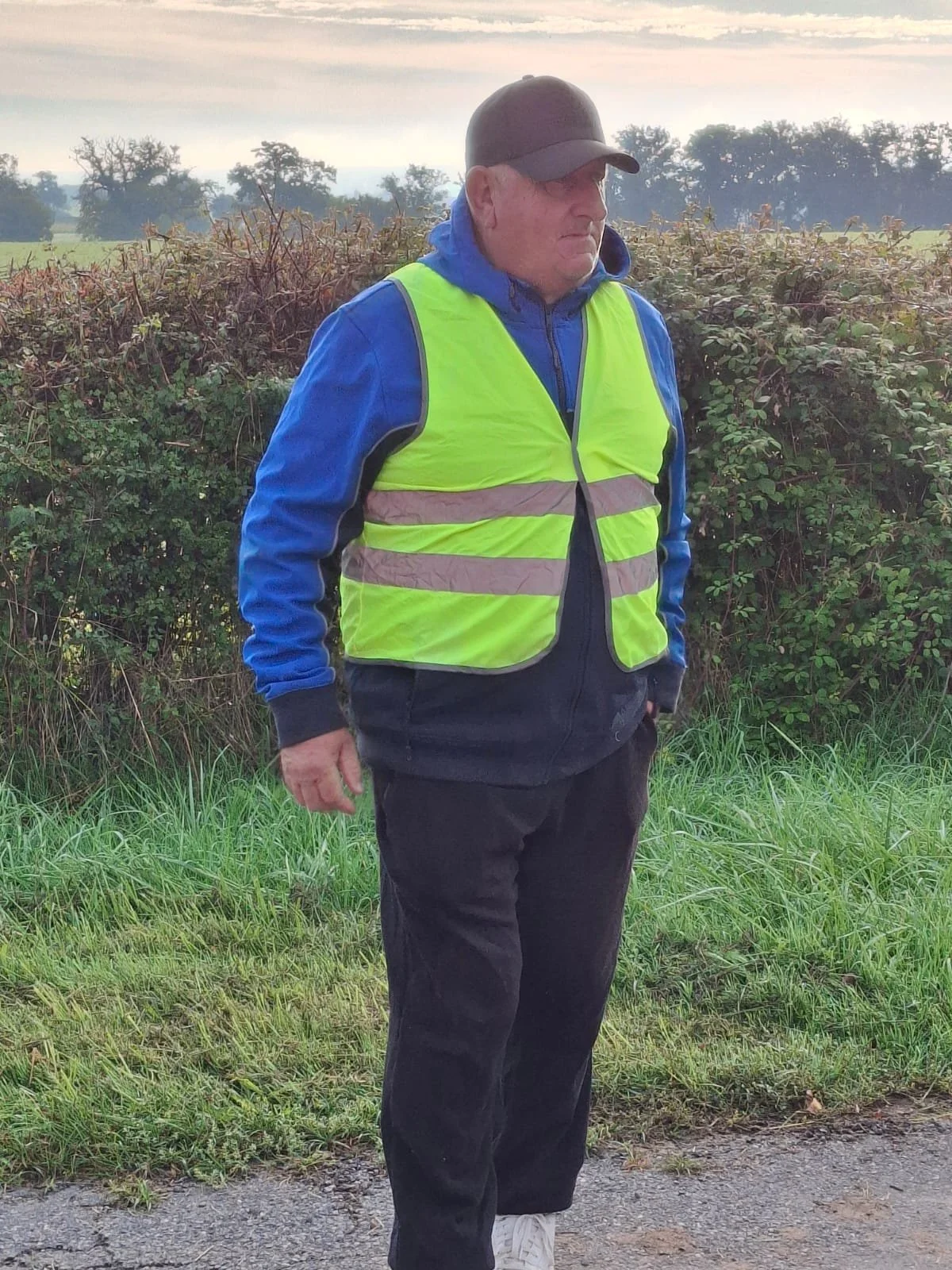 Homme portant un gilet de haute visibilité jaune, une veste bleue, un pantalon noir, un casque noir, et des chaussures blanches, debout à côté d'une haie en plein air, avec un ciel nuageux en arrière-plan.