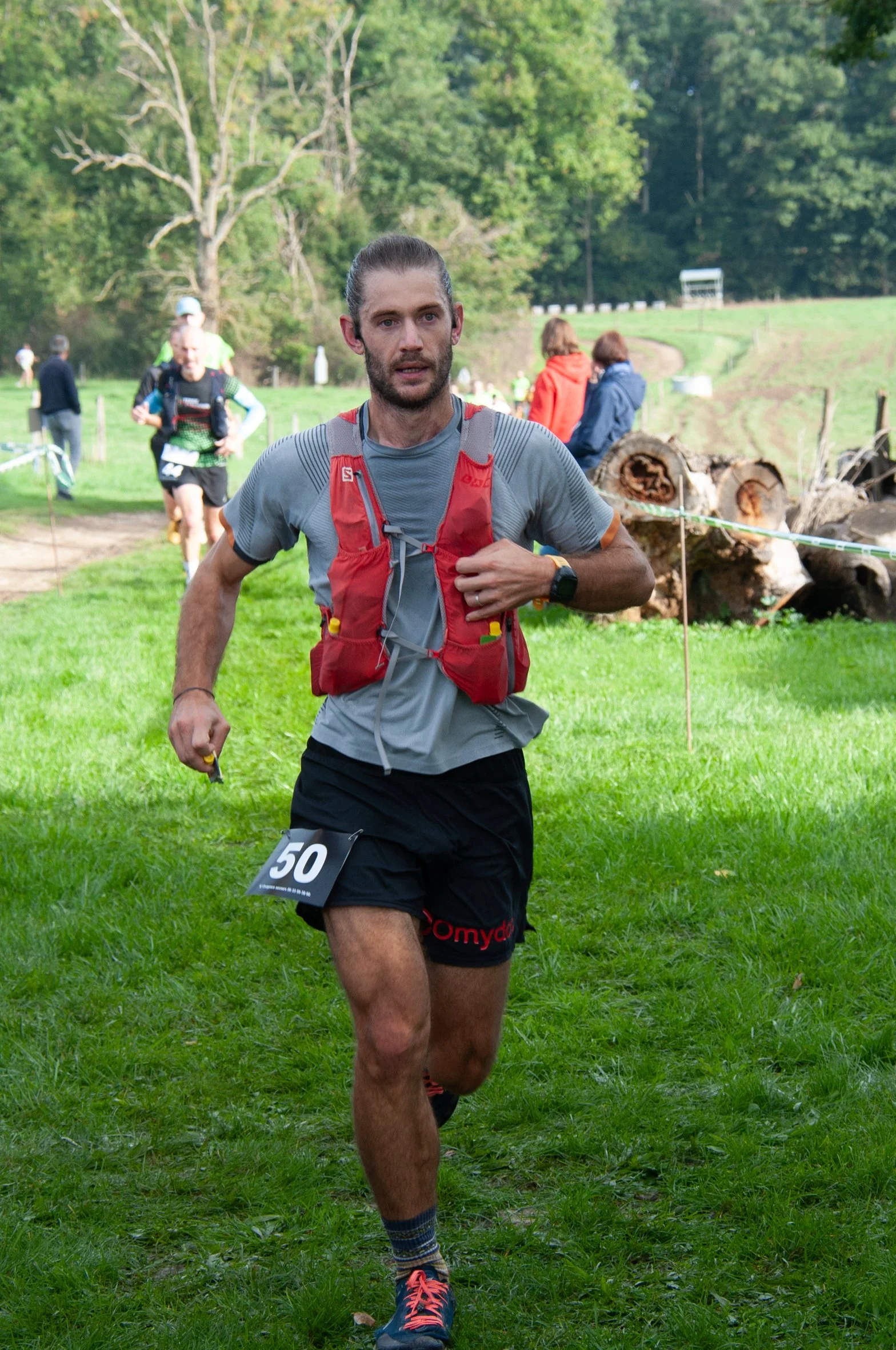 Un homme court lors d'une course en plein air, portant un gilet rouge et un t-shirt gris, avec un numéro 50 attaché à sa jambe gauche. Le fond montre un terrain vert avec quelques spectateurs, des arbres et des bûches coupées.