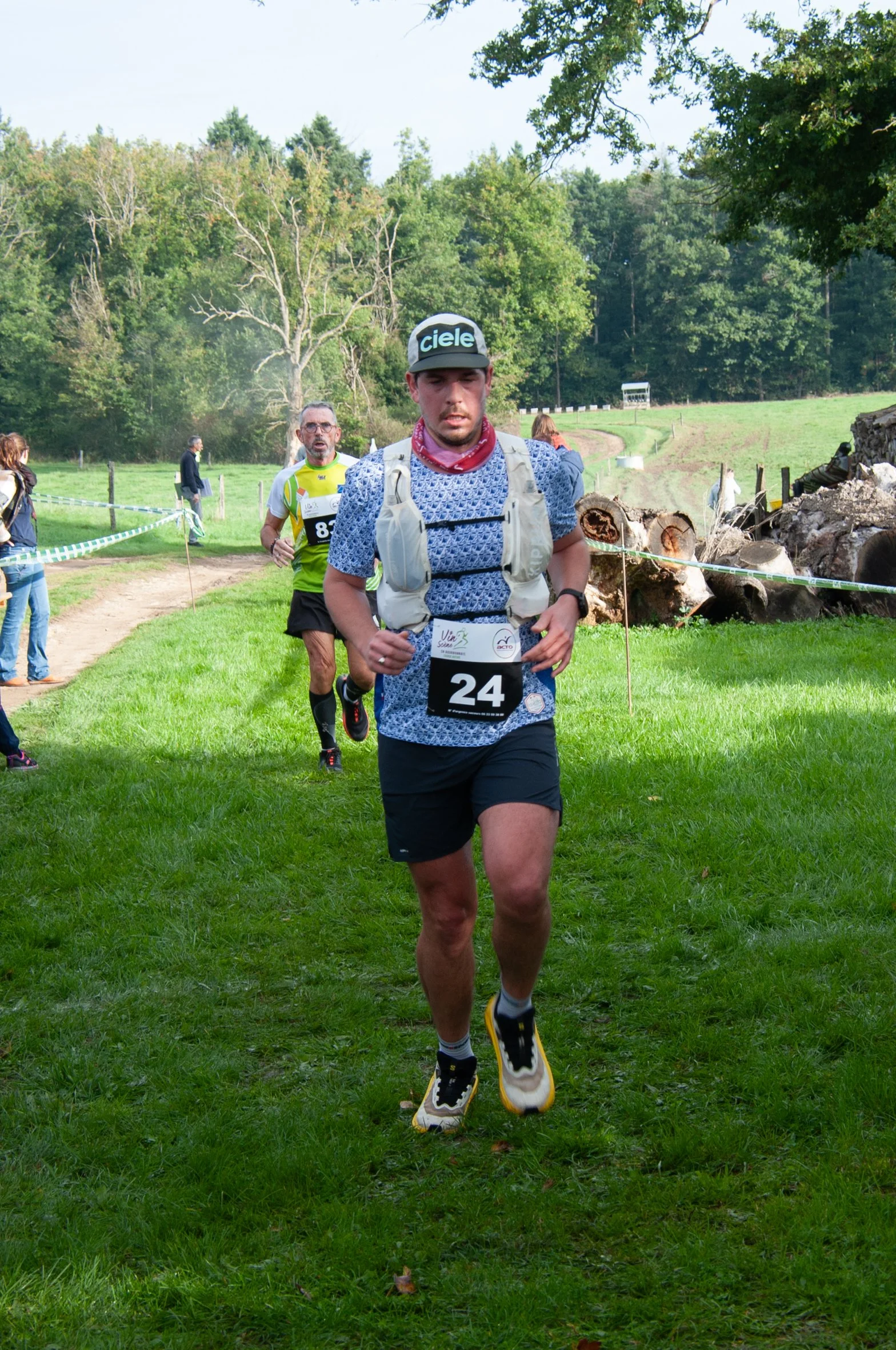 Un homme court lors d'une compétition en plein air, portant un dossard avec le numéro 24, un chapeau noir avec le mot 'ciele', un t-shirt bleu à motifs, un sac à dos gris, et des chaussures de course. Il y a d'autres coureurs en arrière-plan et le pa