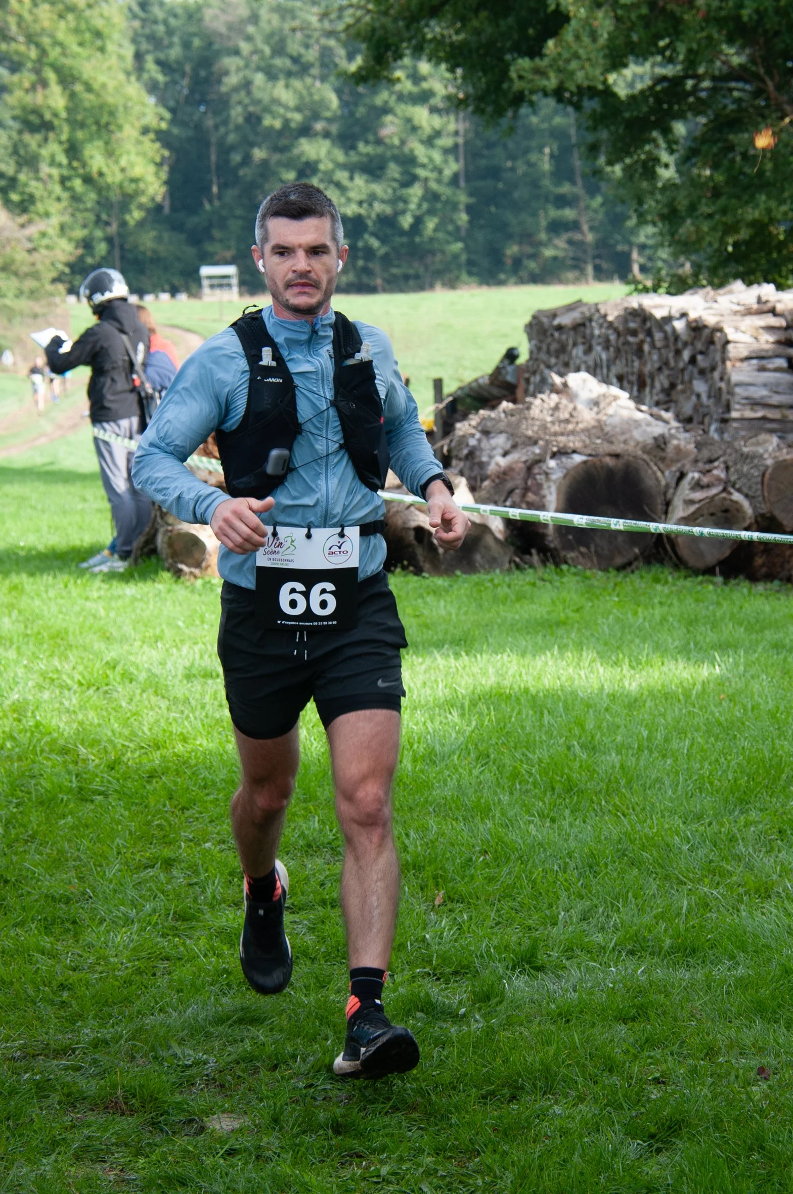 Homme courant lors d'une course dans un environnement naturel avec arbres et bûches, portant un numéro 66, une veste légère et des écouteurs.