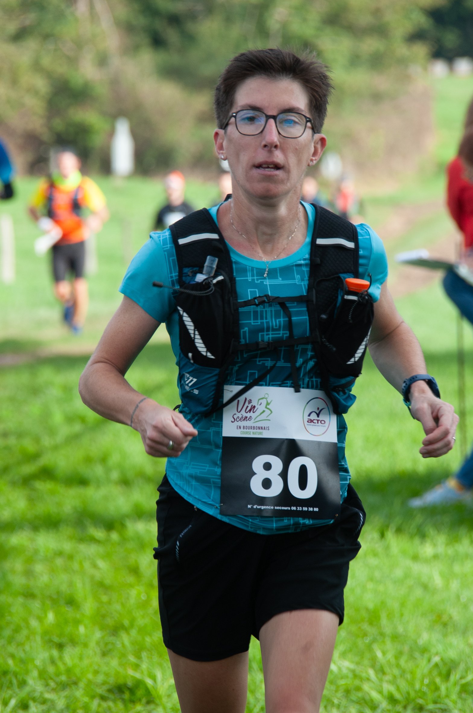 Femme courant lors d'une course en extérieur, portant un numéro 80, une montre et un sac à dos, avec un environnement vert et des autres coureurs en arrière-plan.