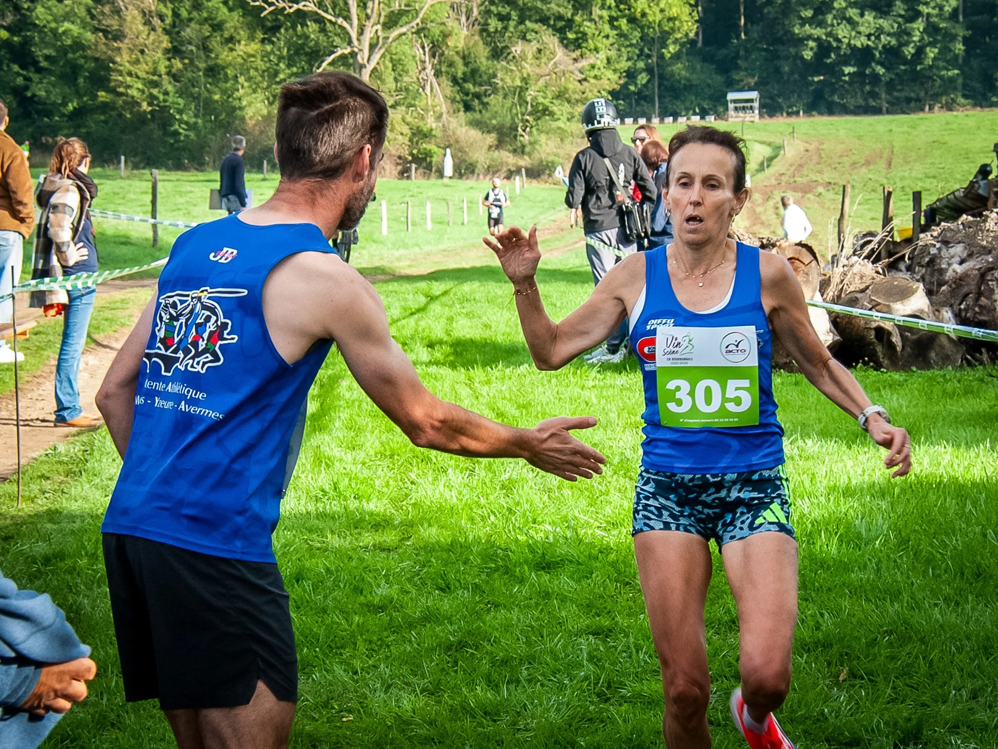 Une femme en course, portant un dossard vert avec le numéro 305, échange un coup de main avec un homme en vestiaire bleu lors d'une compétition en plein air.