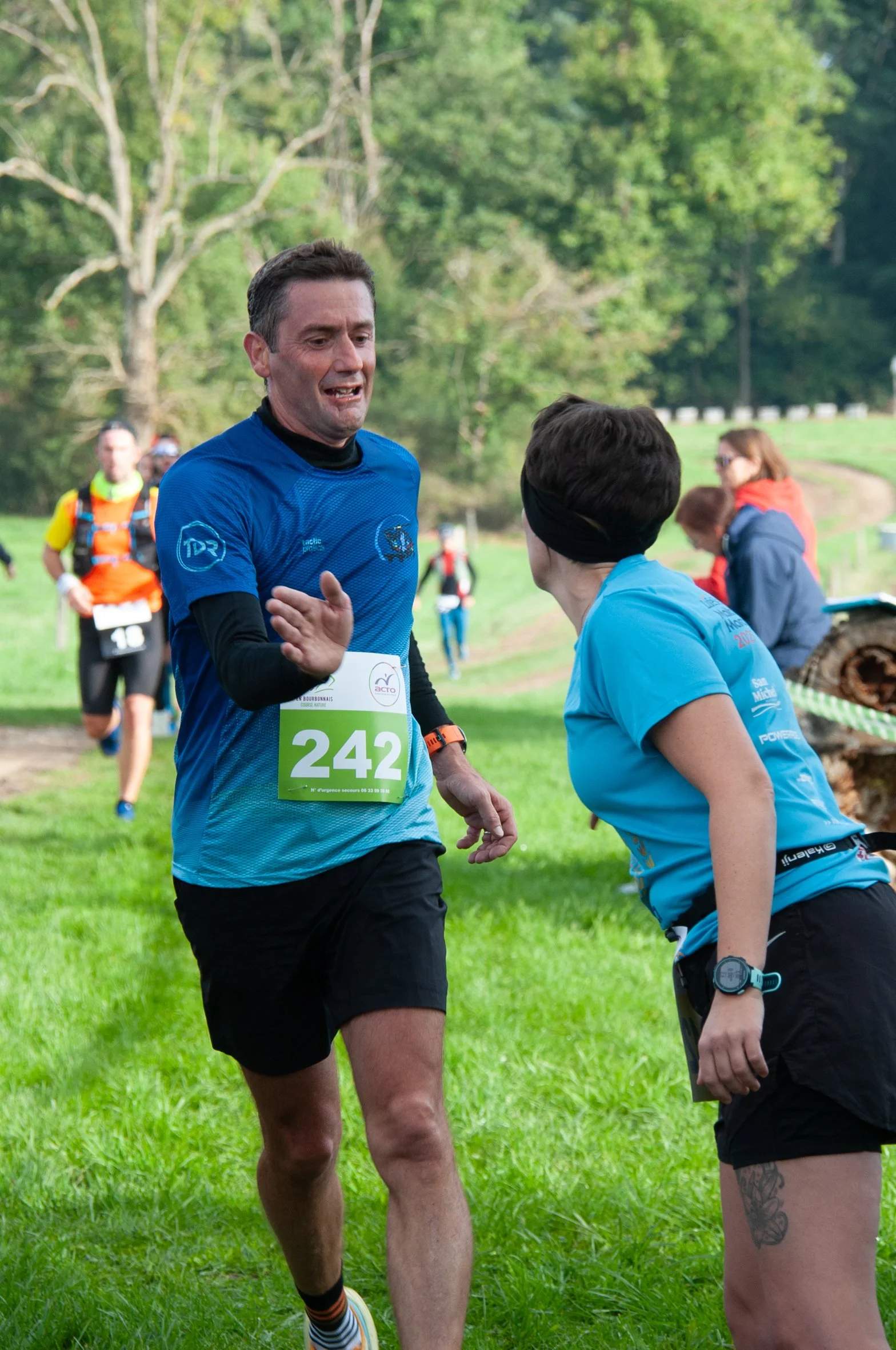 Deux coureurs échangeant un high-five lors d'une course en plein air, avec d'autres coureurs en arrière-plan sur une pelouse verte dans un parc.