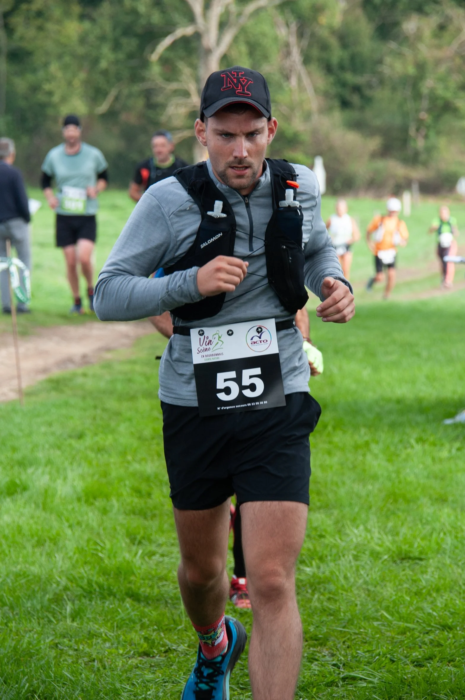 Un homme court lors d'une course en plein air, portant un dossard numéro 55, un bonnet noir avec logo rouge N Y, une veste grise et un sac à dos, avec d'autres coureurs en arrière-plan dans un cadre naturel.