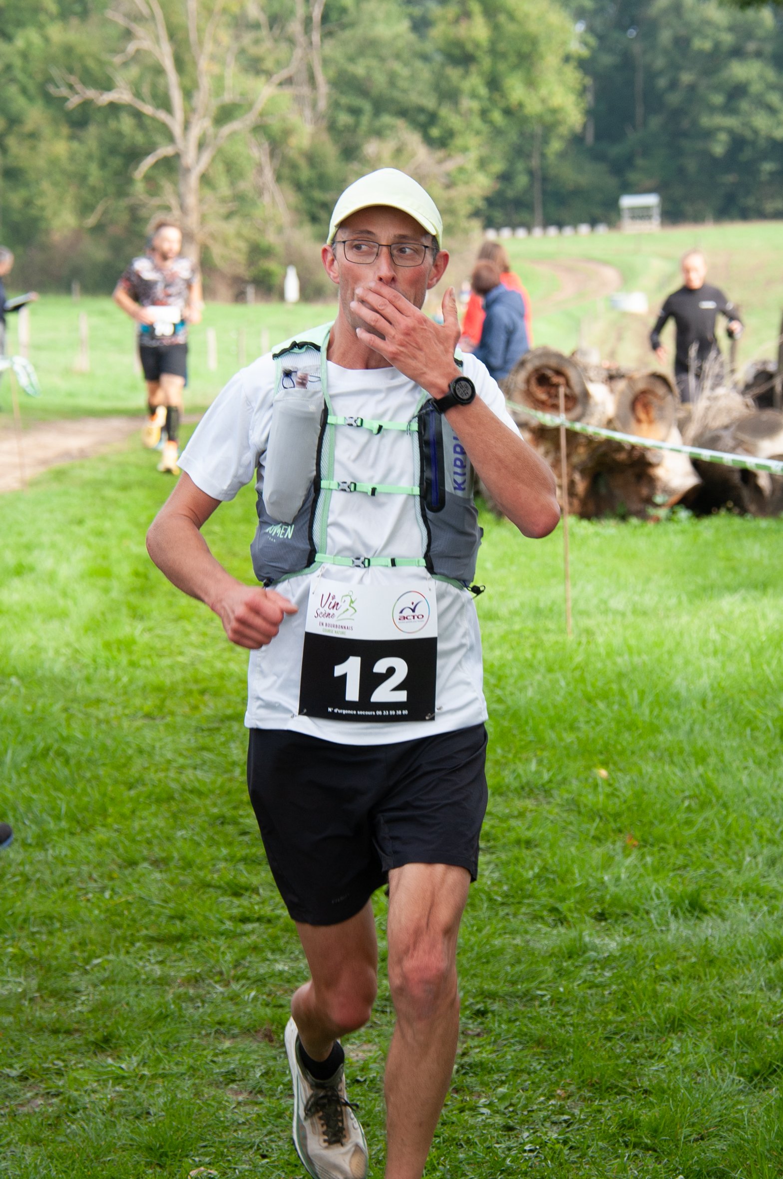 Un homme participant à une course en extérieur, portant un t-shirt blanc, un short noir, un chapeau clair, et un sac d'hydratation, avec le numéro 12, en train de courir ou marcher sur un chemin herbeux.