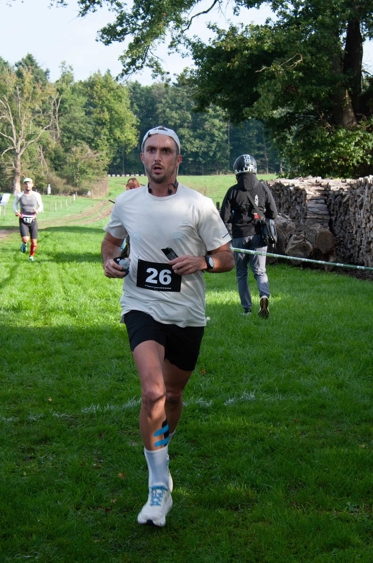 Un homme court lors d'une course en pleine nature, portant un maillot blanc, un short noir, une casquette blanche et un dossard avec le numéro 26.