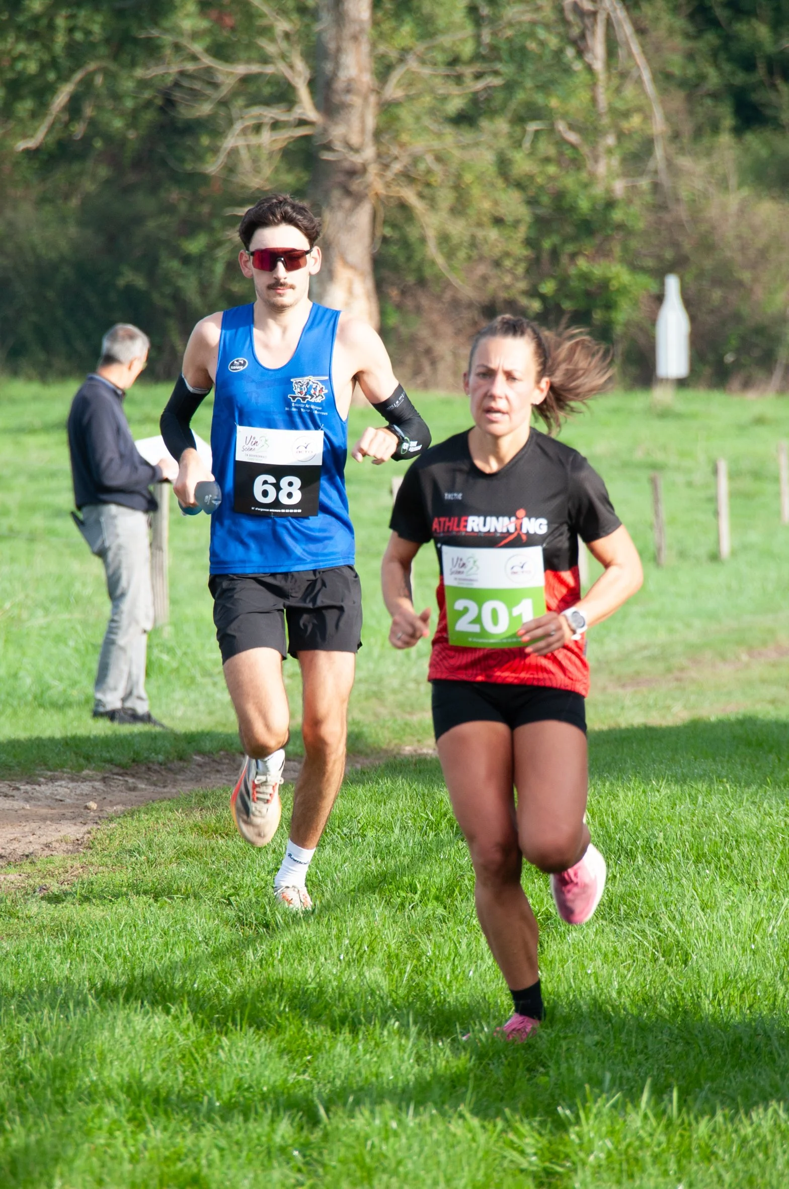 Deux coureurs, un homme avec un maillot bleu et une femme avec un t-shirt noir et rouge, participent à une course dans un parc vert. Ils portent des numéros 68 et 201.