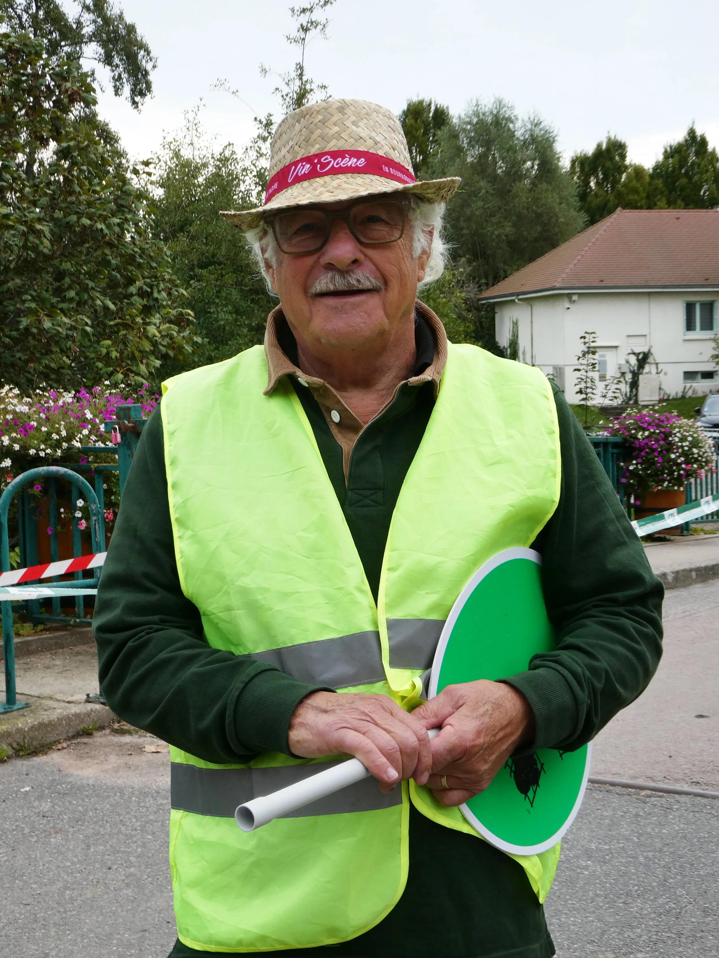 Homme âgé portant un chapeau en paille avec un ruban rose, une veste jaune de sécurité, tenant un panneau vert avec une image de coccinelle, dans un environnement résidentiel avec des maisons et des plantes.