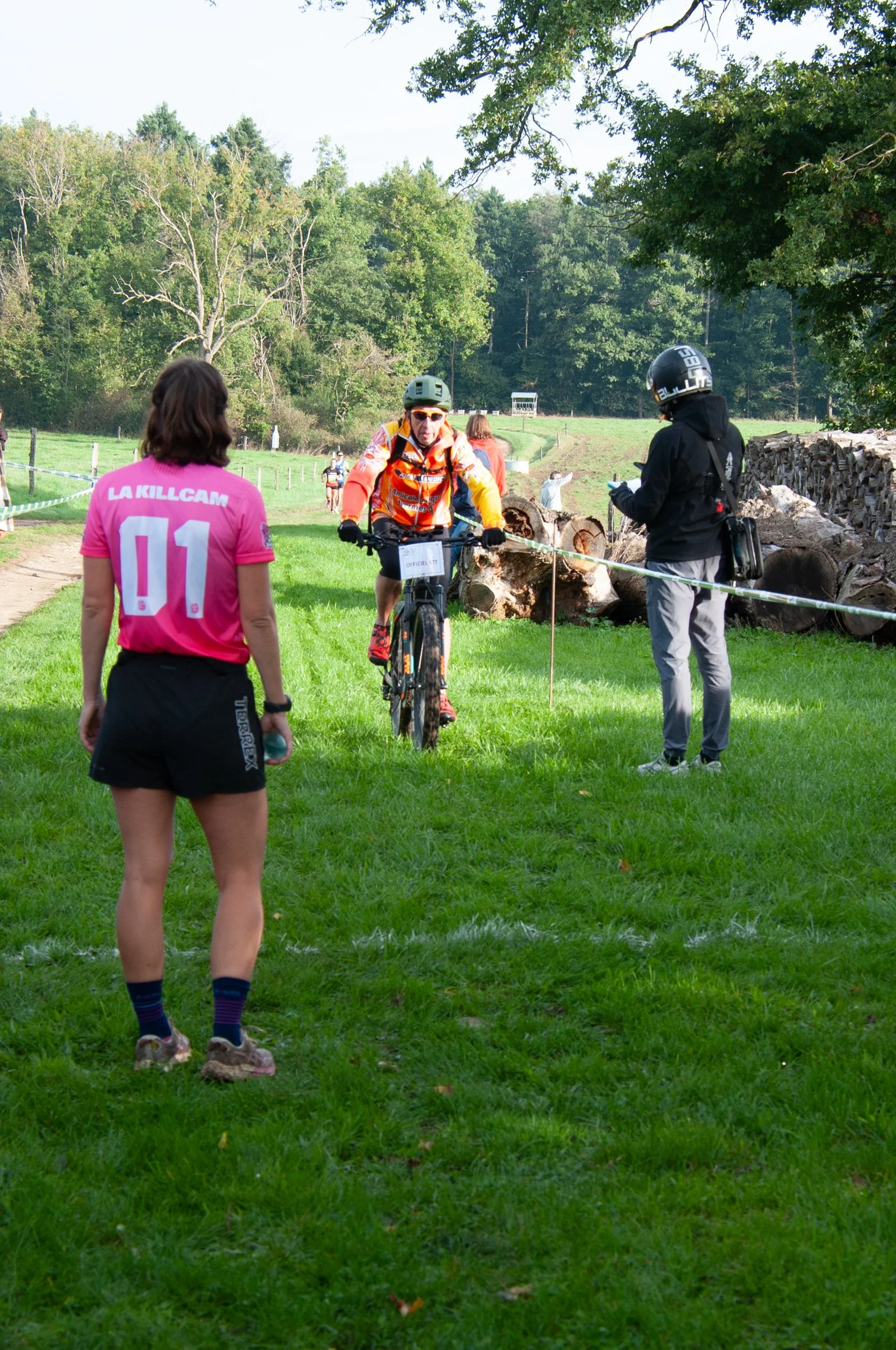 Un coureur de vélo passé au contrôle d'un événement sportif, avec une femme en t-shirt rose et un homme en casque noire, dans un environnement extérieur verdoyant.