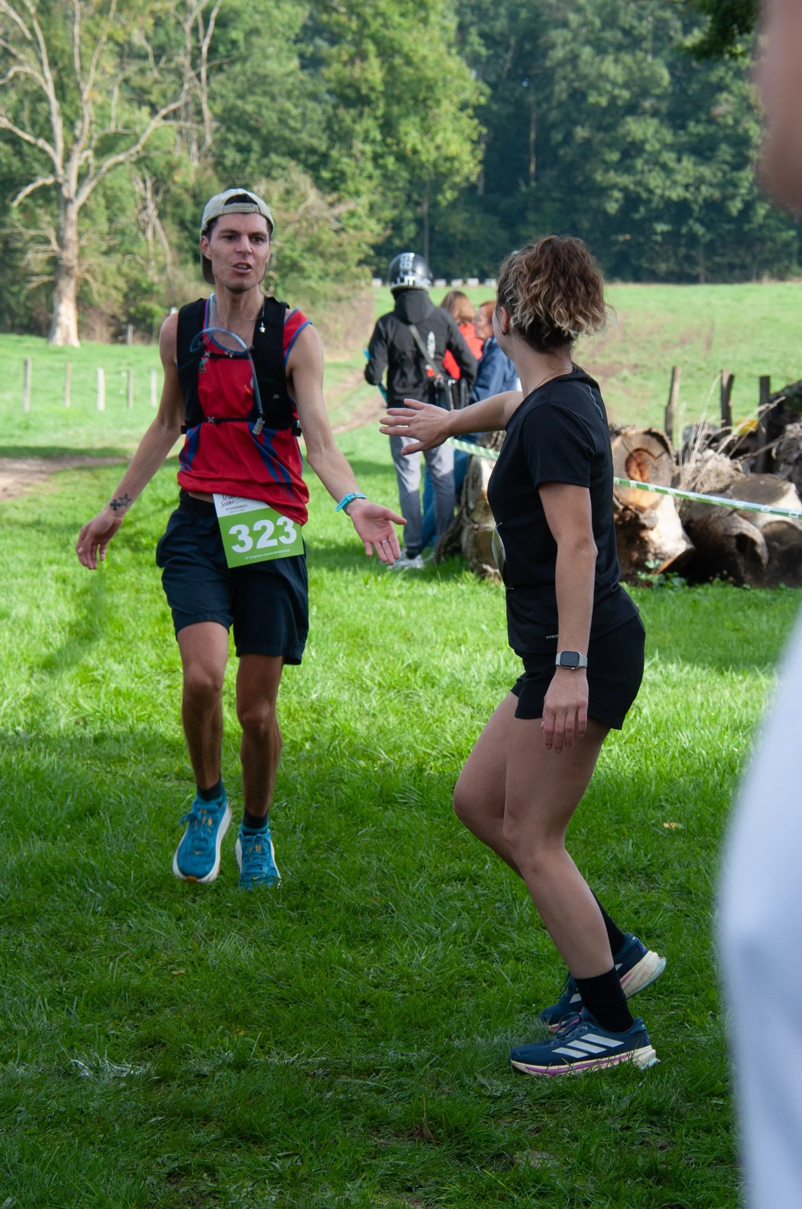 Un homme et une femme discutent lors d'un événement sportif en plein air, avec l'homme portant un dossard numéro 323 et une casquette, et la femme gesticulant vers lui dans un environnement verdoyant.
