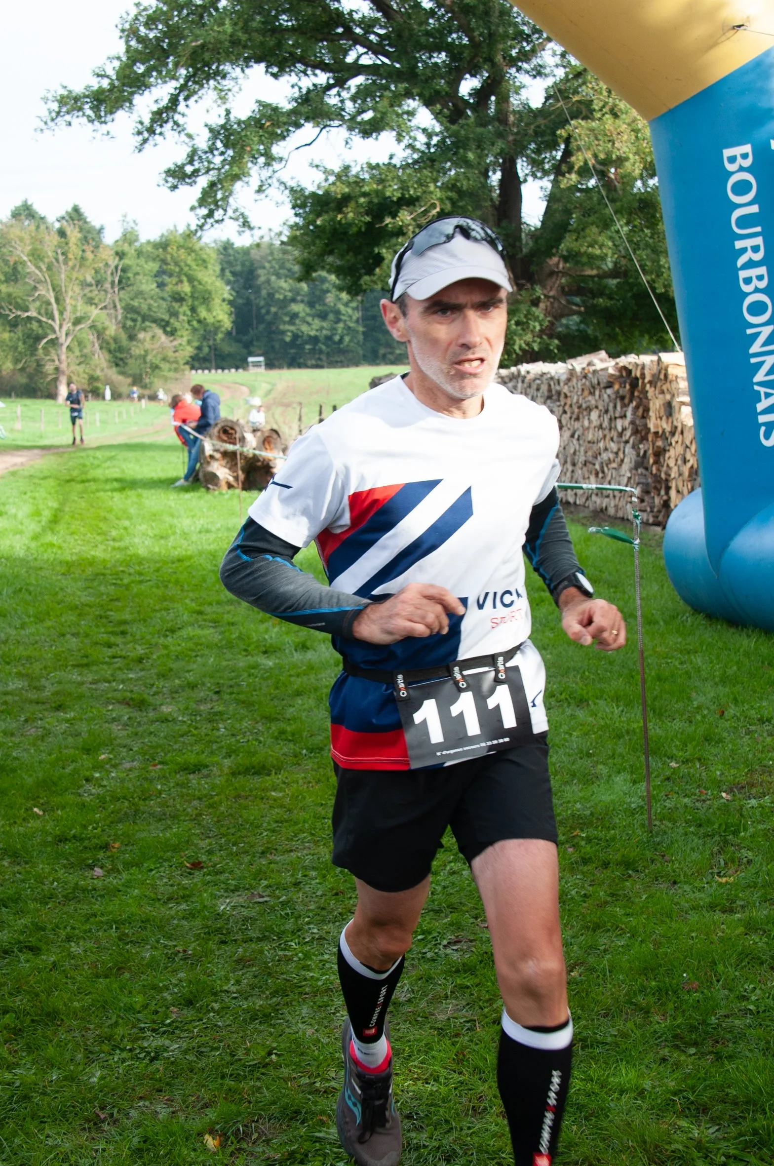 Un homme court lors d'une course de trail dans un espace vert, portant un maillot blanc avec des rayures bleues et rouges, un casque blanc, un bracelet de montre connectée, et un dossard avec le numéro 111.