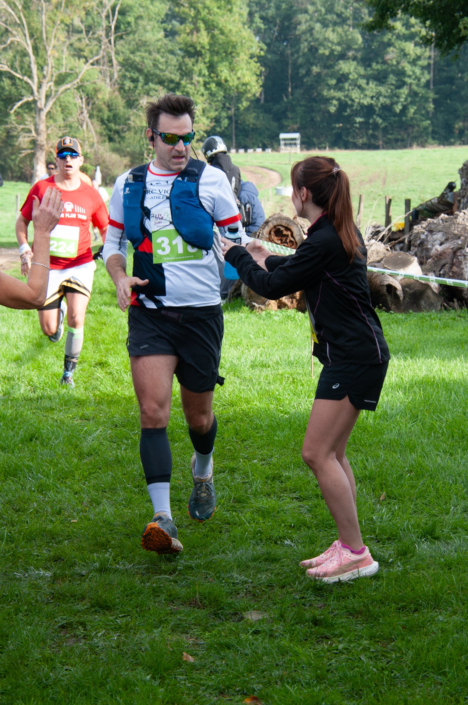 Un coureur remet un objet à une bénévole lors d'une course à pied en plein air, avec d'autres coureurs en arrière-plan.