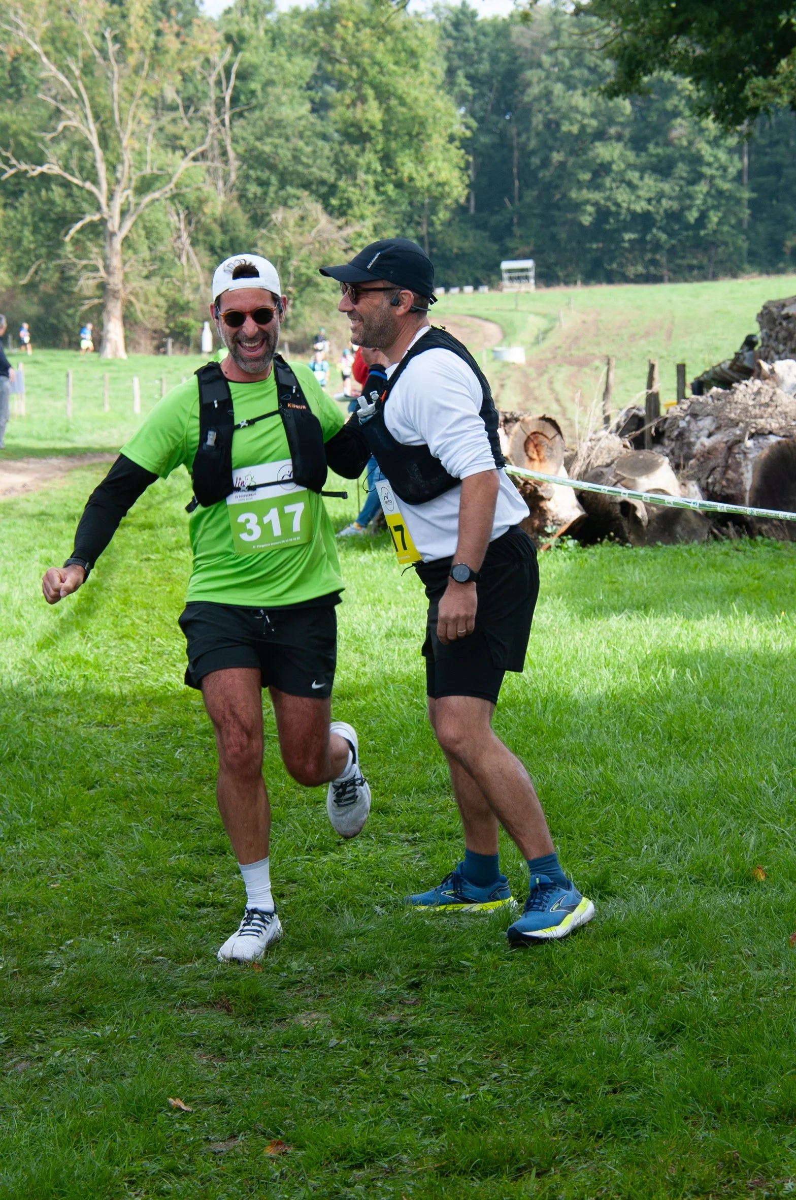 Deux hommes souriants en tenue de course à pied, échangeant un moment de joie lors d'une compétition en plein air, avec un environnement verdoyant et arbres en arrière-plan.