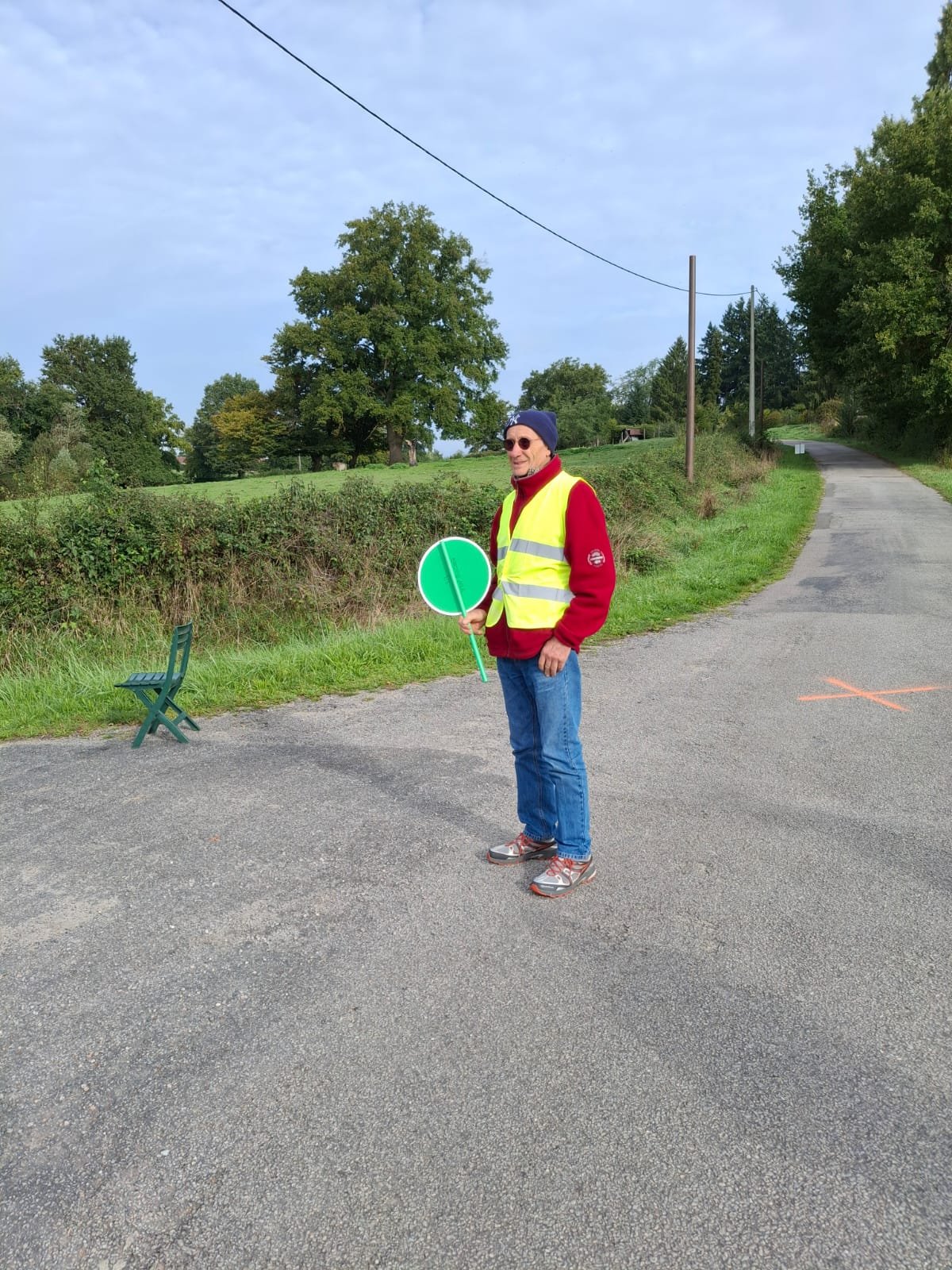 Un homme portant une veste rouge, un gilet jaune fluorescent, un bonnet et des lunettes de soleil, tient un panneau de signalisation vert dans une campagne. Il se tient sur une route pavée avec des arbres et un ciel partiellement nuageux en arrière-p