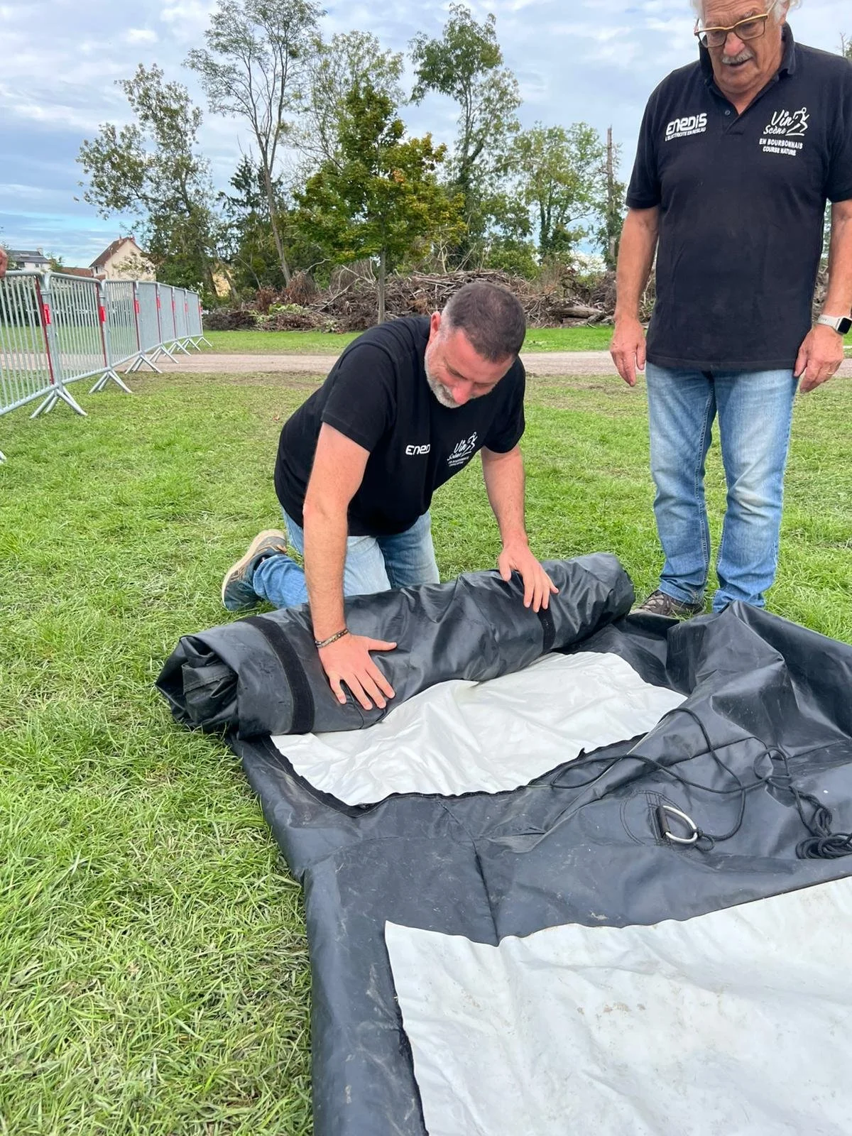 Deux hommes déballent un tapis de camping ou de secours sur une zone herbeuse, avec des barrières métalliques en arrière-plan et des arbres.