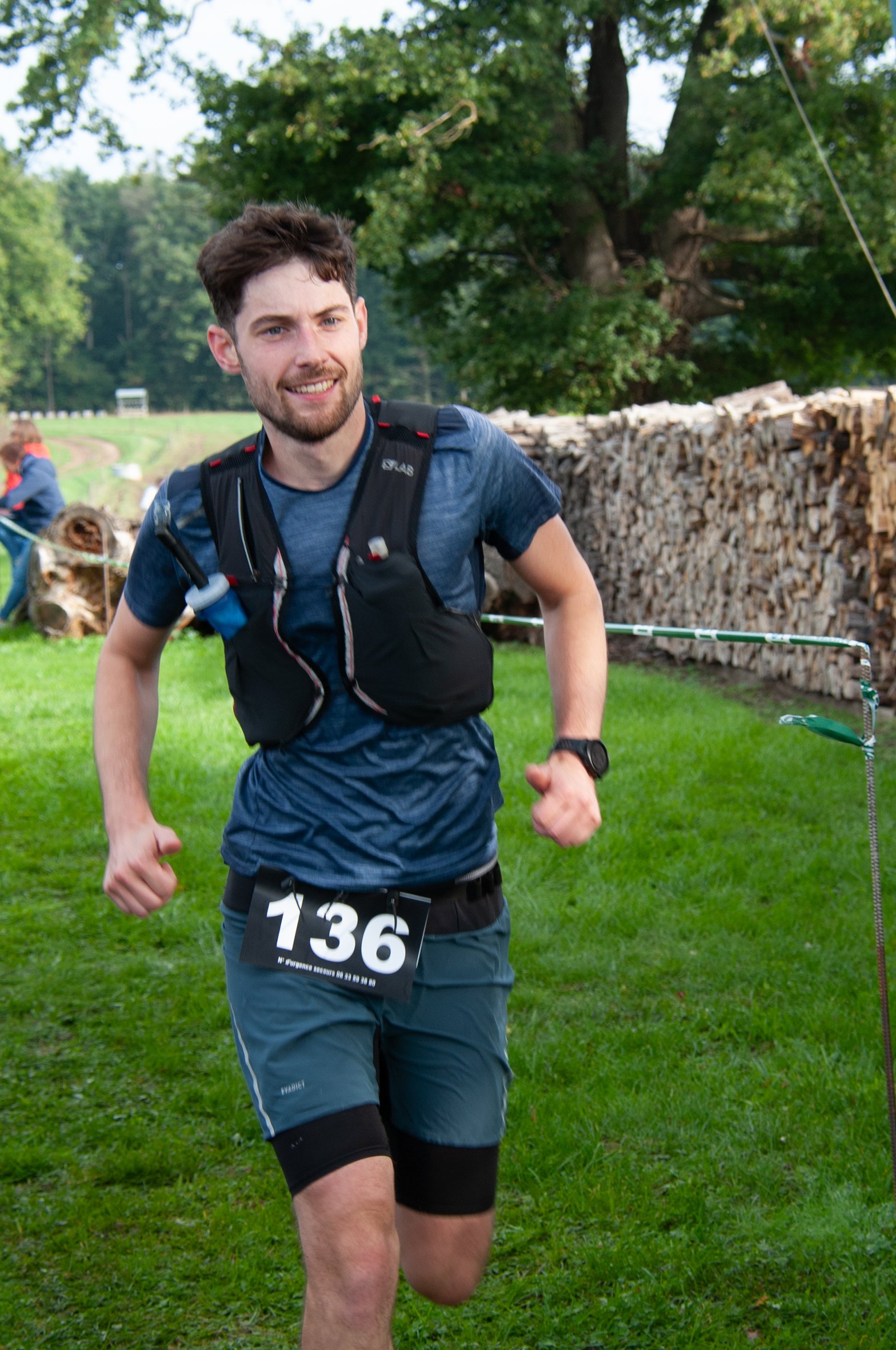 Un homme court lors d'une course en extérieur, portant un numéro 136, vêtu d'une tenue de course et d'un gilet avec des bouteilles d'eau.