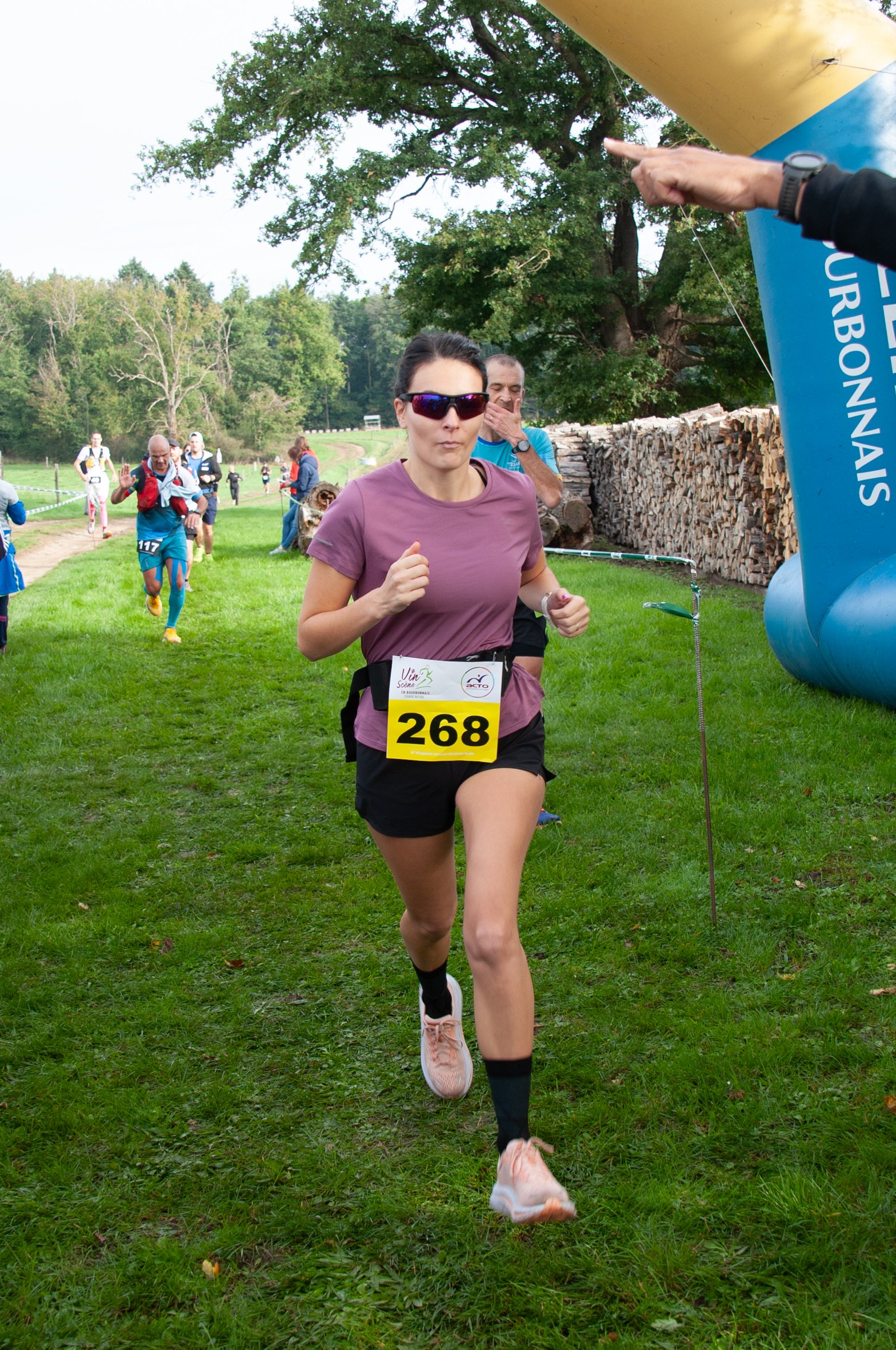 Une femme court lors d'une course en extérieur, portant un dossard numéro 268, lunettes de soleil, t-shirt violet, short noir et chaussures de course. D'autres coureurs courent derrière elle. Il y a des arbres verts et un pile de bois en arrière-plan