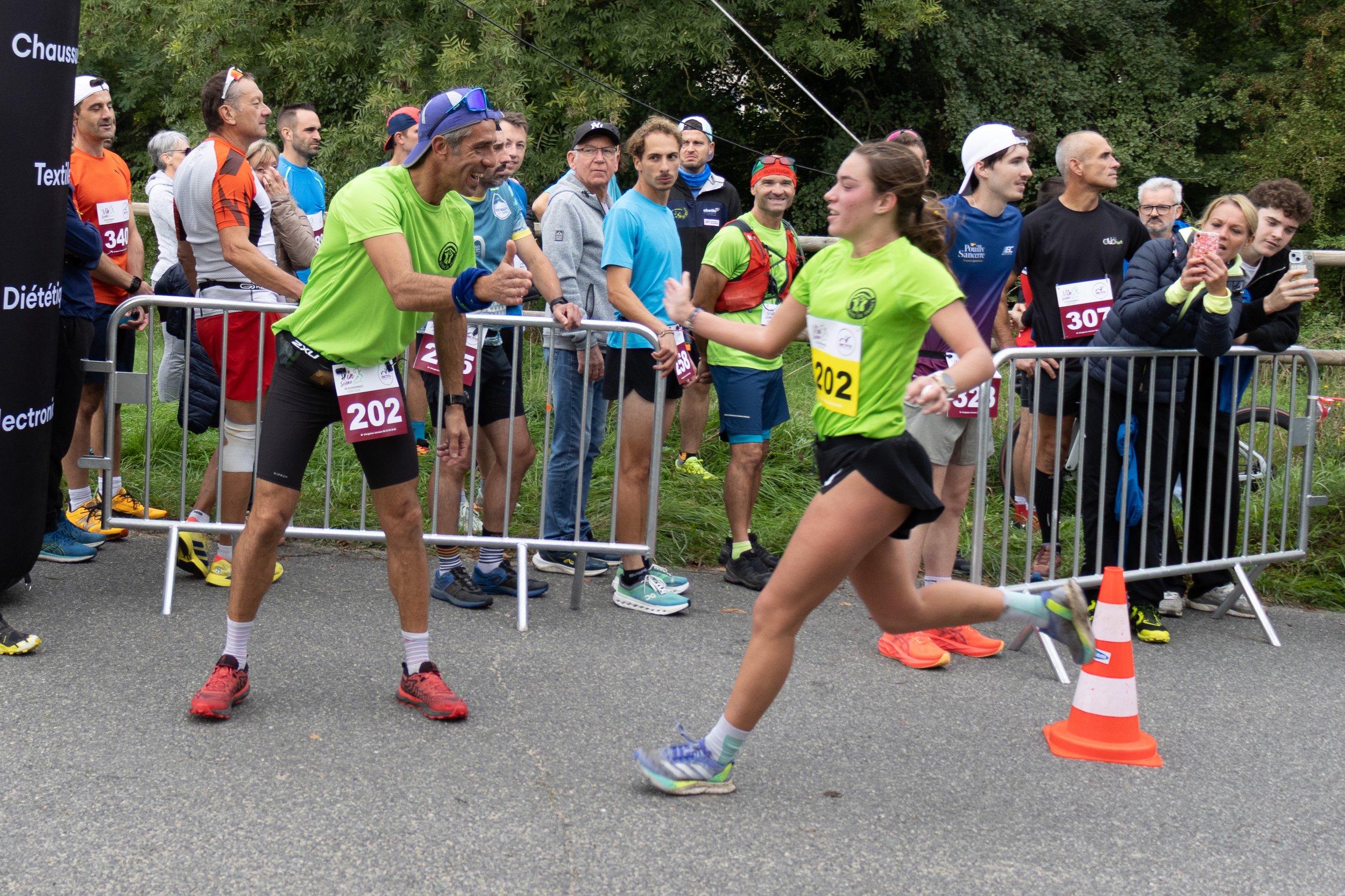 Une femme en tenue de course à pied accélère pour dépasser un homme lors d'une course, avec des spectateurs applaudissant et prenant des photos derrière une barrière.