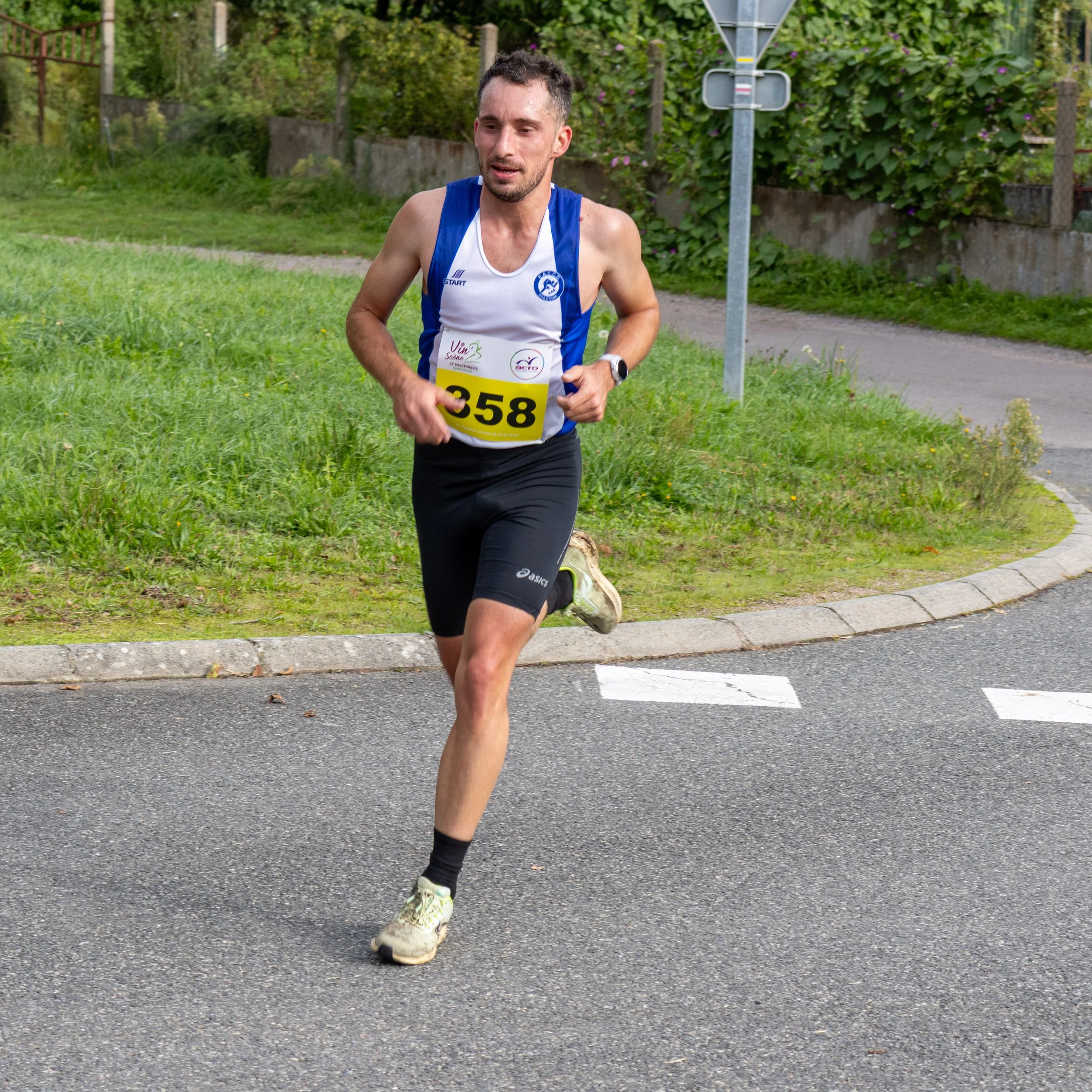 Un homme court lors d'une course, portant un maillot blanc et bleu avec un dossard jaune portant le numéro 358, sur une route avec du verglas, entouré de verdure.