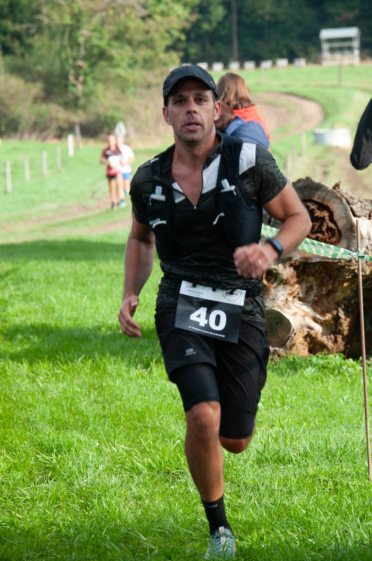 Un homme court lors d'une course en extérieur, portant un dossard avec le numéro 40, un chapeau noir, une montre connectée, un t-shirt noir et des shorts noirs. Il y a des arbres et des autres coureurs en arrière-plan.