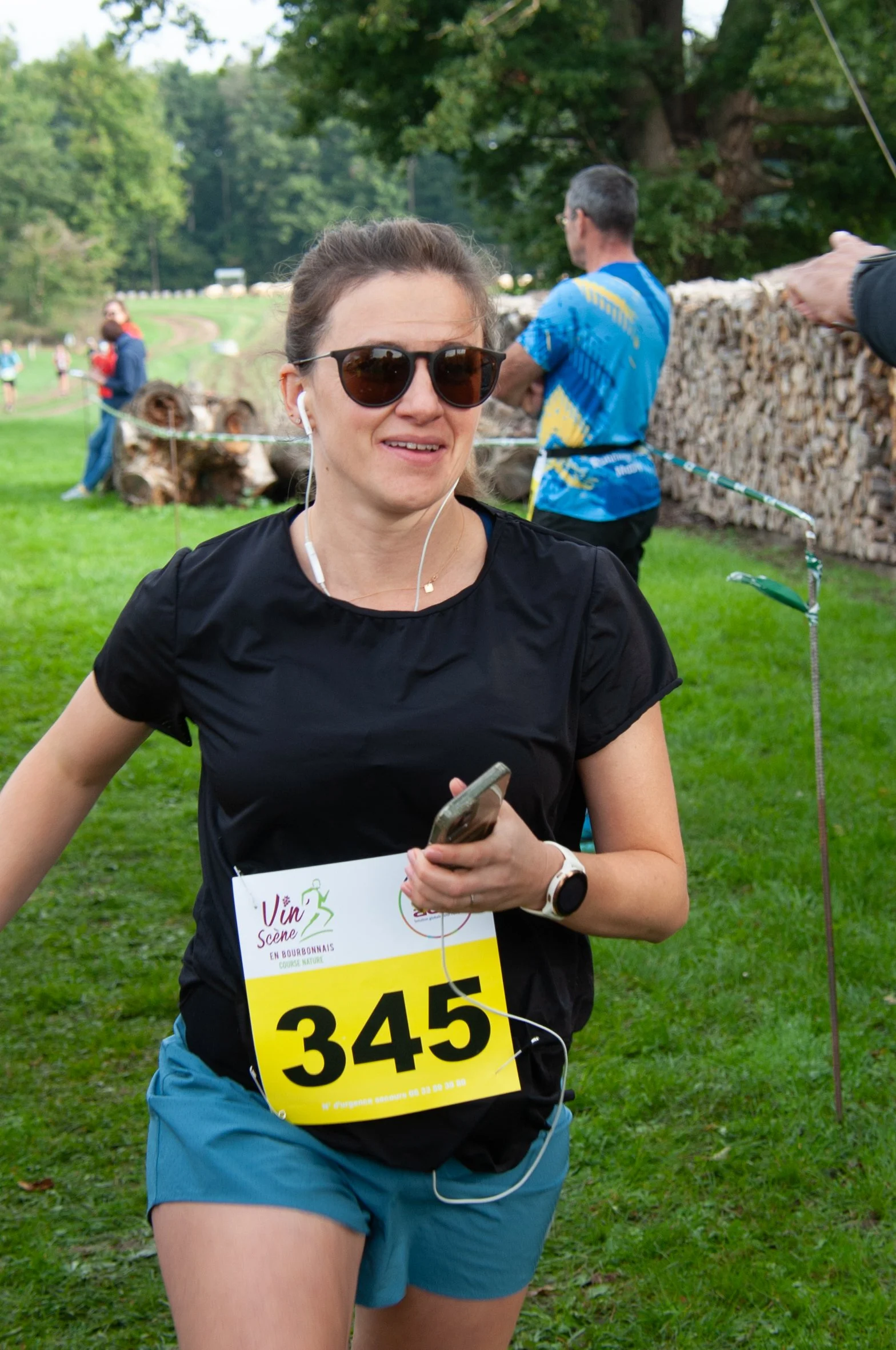 Une femme courait lors d'une course, portant un t-shirt noir, un short bleu, des lunettes de soleil, un écouteur, un montre connectée, et un numéro de course 345. Le fond montre une zone verte avec des arbres, un enclos en bois, et d'autres participa