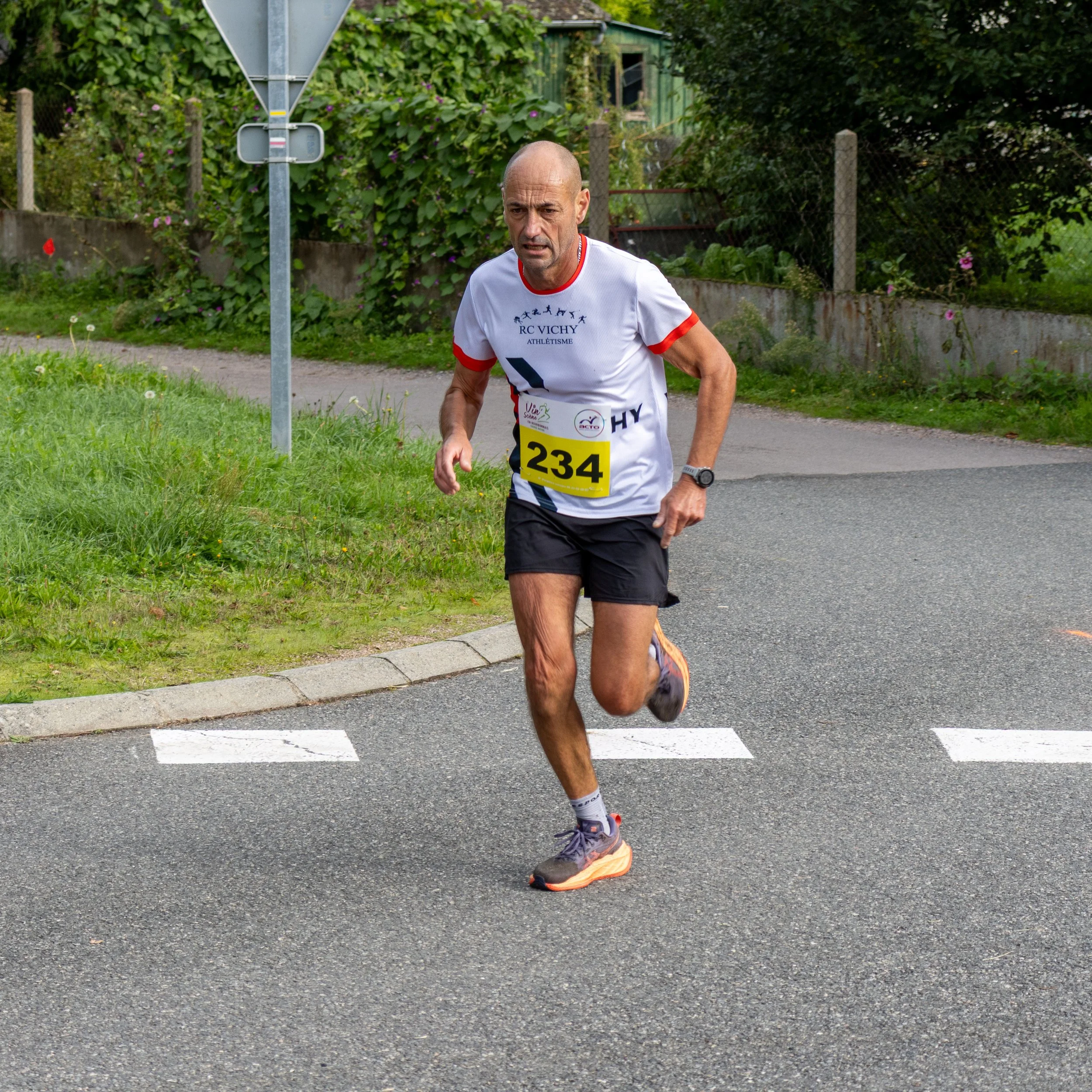 Un homme court sur une route pavée lors d'une course, portant un t-shirt blanc avec des textes et un numéro 234, et des chaussures de course.
