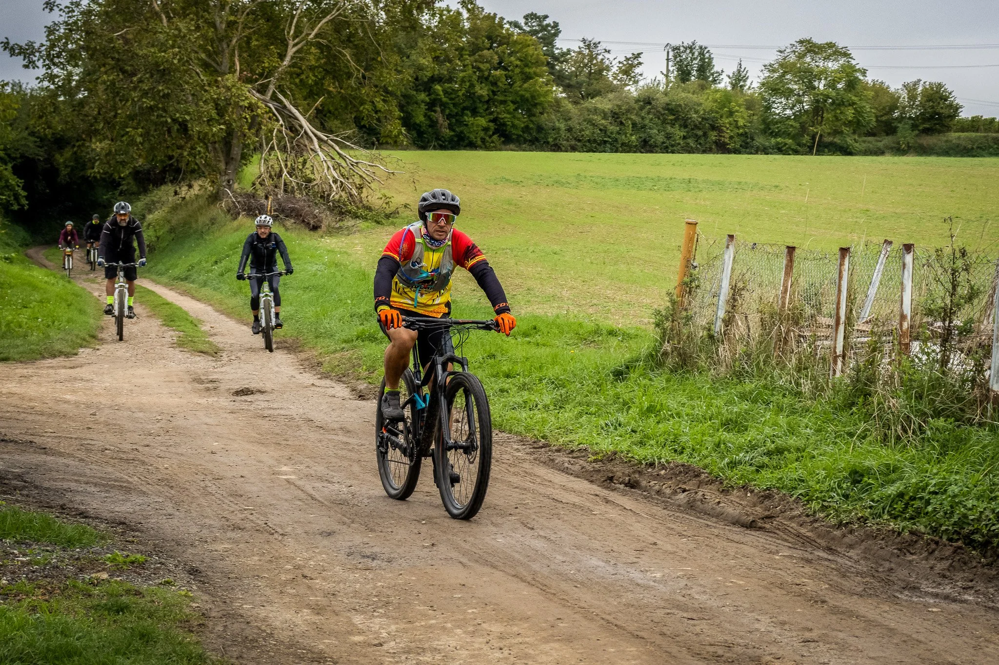 Groupe de cyclistes pédalant sur un chemin de terre au milieu de la campagne, avec arbres et champs verts environnants.