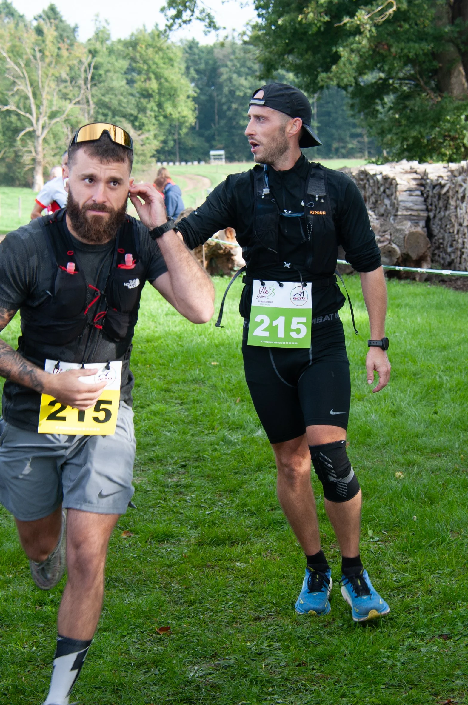 Deux hommes en tenue de course à pied lors d'une compétition en plein air, l'un avec un numéro 215 et l'autre lui donnant des conseils ou de l'encouragement, avec un environnement naturel verdoyant en arrière-plan.