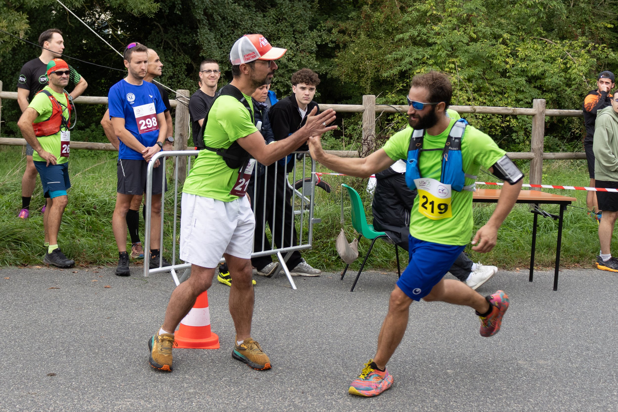 Deux coureurs de marathon se félicitant avec une poignée de main à la fin de la course, entourés d'autres participants et spectateurs dans un cadre extérieur vert.