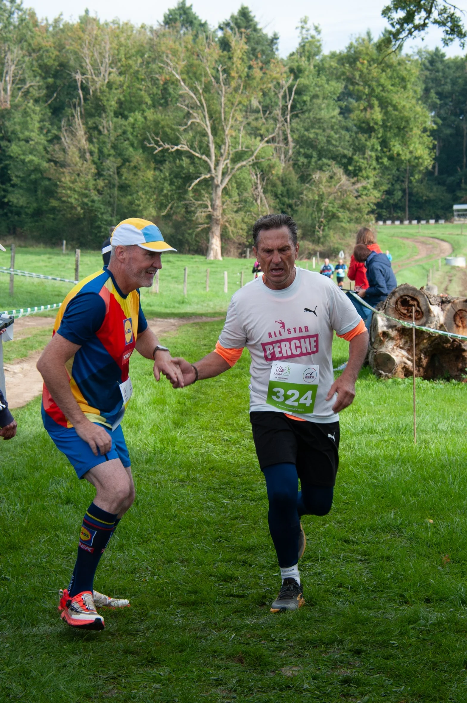 Deux coureurs masculins se serrent la main dans un environnement naturel lors d'une course, avec des arbres et un chemin en arrière-plan.