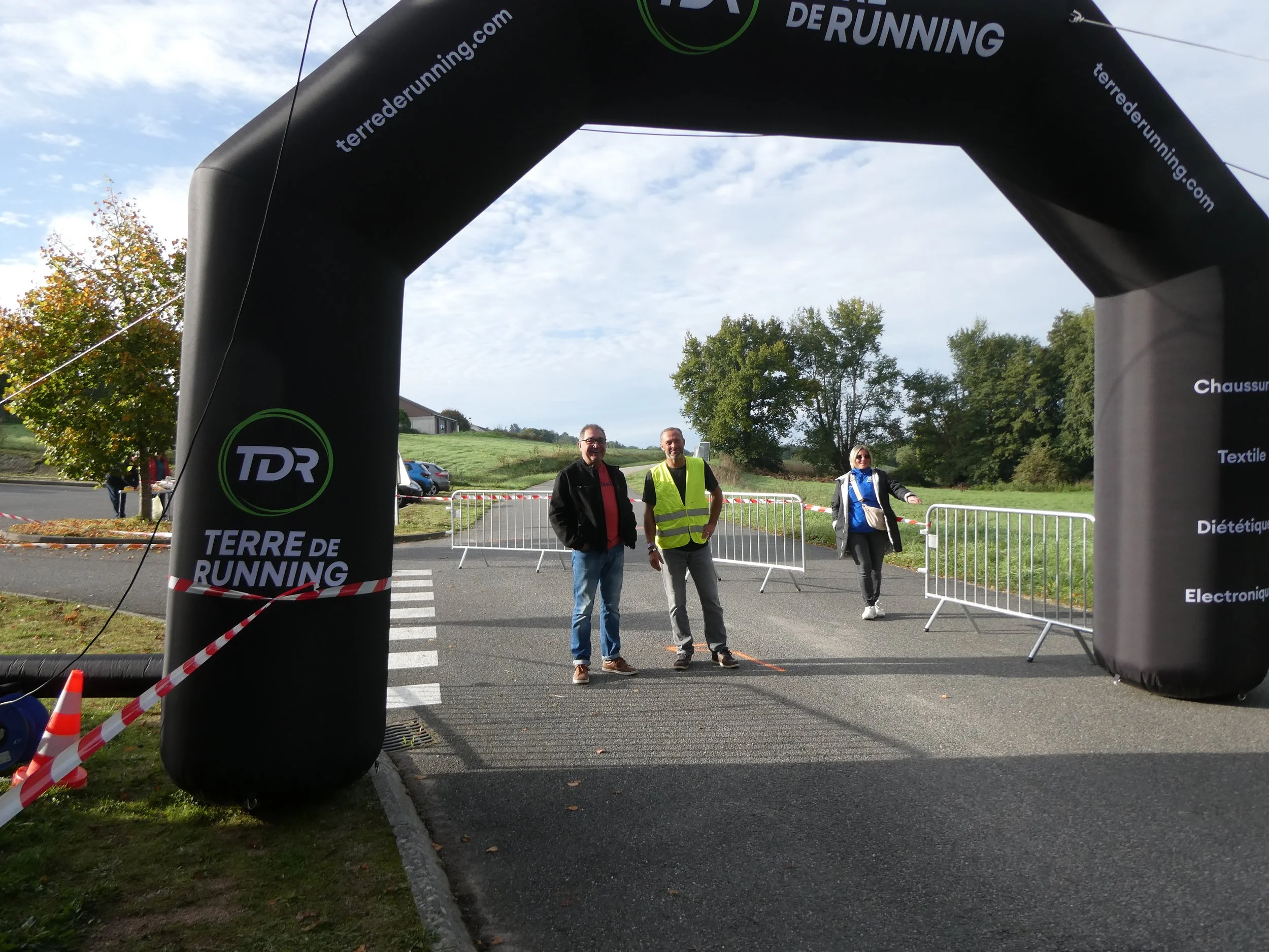 Trois personnes se tiennent sous une arche gonflable noire portant le logo TDR TERRE DE RUNNING, avec un terrain et des arbres en arrière-plan, lors d'un événement de course à pied.