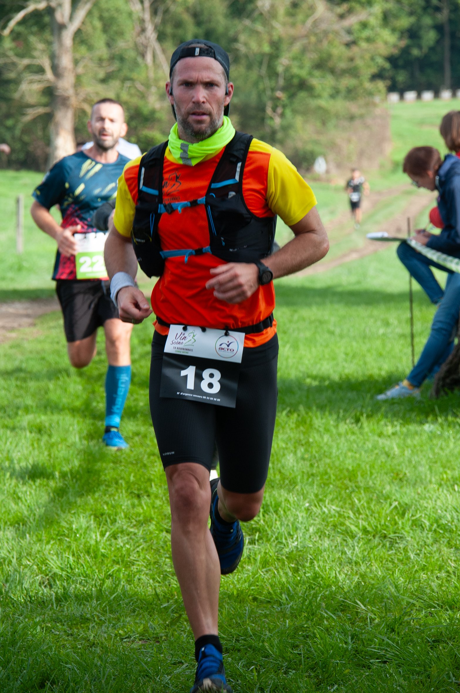 Un homme court lors d'une course en extérieur, portant un maillot orange et jaune, un casque noir, un porte-bidon, un bracelet de montre, et un dossard numéro 18, avec d'autres coureurs en arrière-plan dans un cadre naturel.