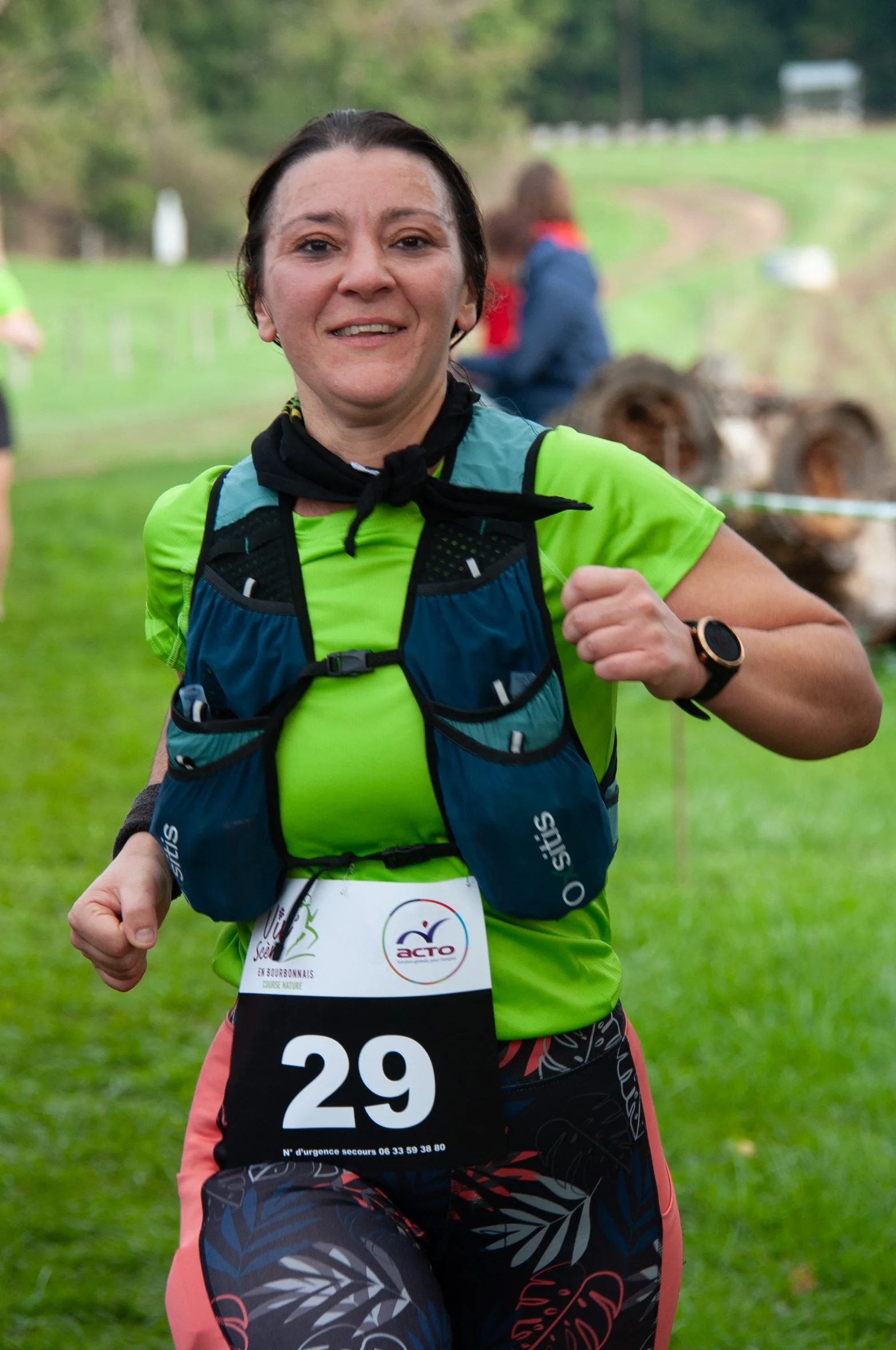 Femme courrant lors d'une course en plein air, portant un numéro 29, un maillot vert, un gilet de course bleu et un bracelet connecté, avec un fond de verdure.