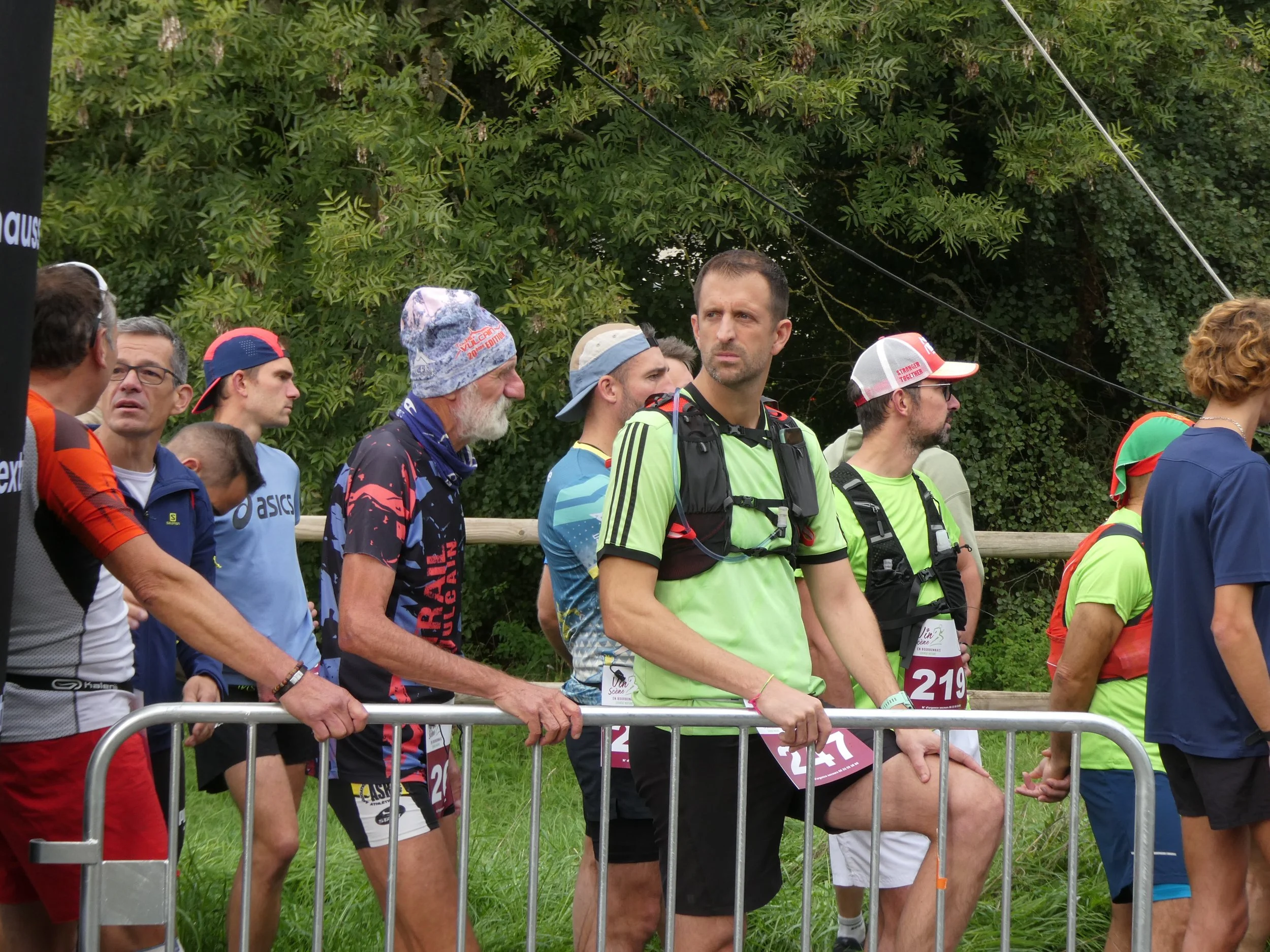 Groupe de coureurs en ligne, prêts pour une course, certains avec numéros sur leur maillot, dans un environnement extérieur avec des arbres en arrière-plan.