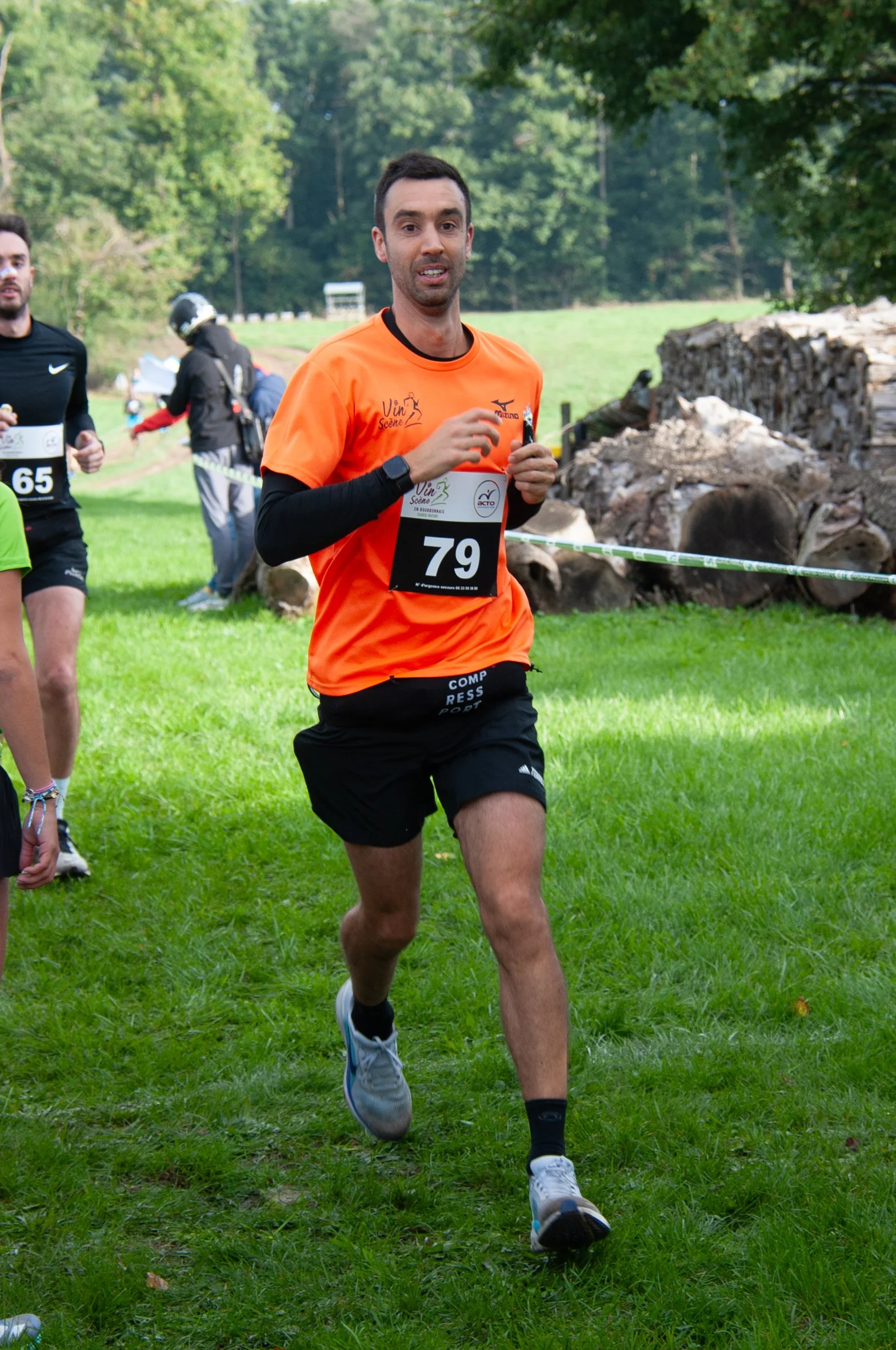 Un homme court lors d'une course en pleine nature, portant un t-shirt orange avec un numéro 79, derrière des piles de bois et des arbres.