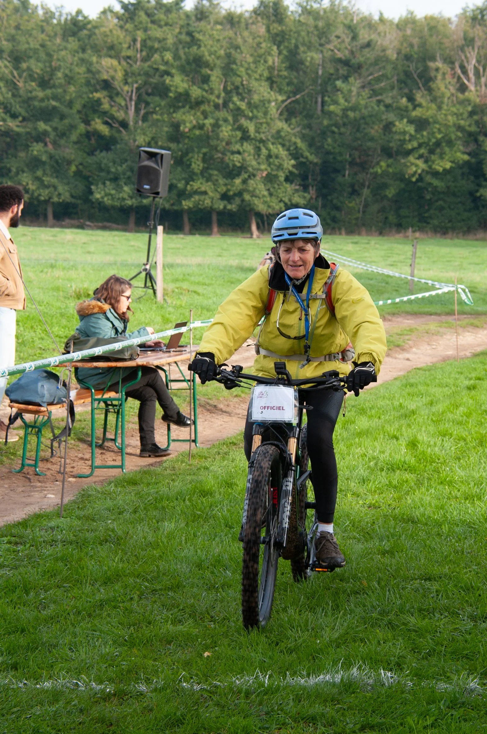 Une femme à vélo portant un casque dans un environnement extérieur verdoyant, avec une équipe de personnes à une table à côté, dans un cadre probablement organisé ou compétitif.