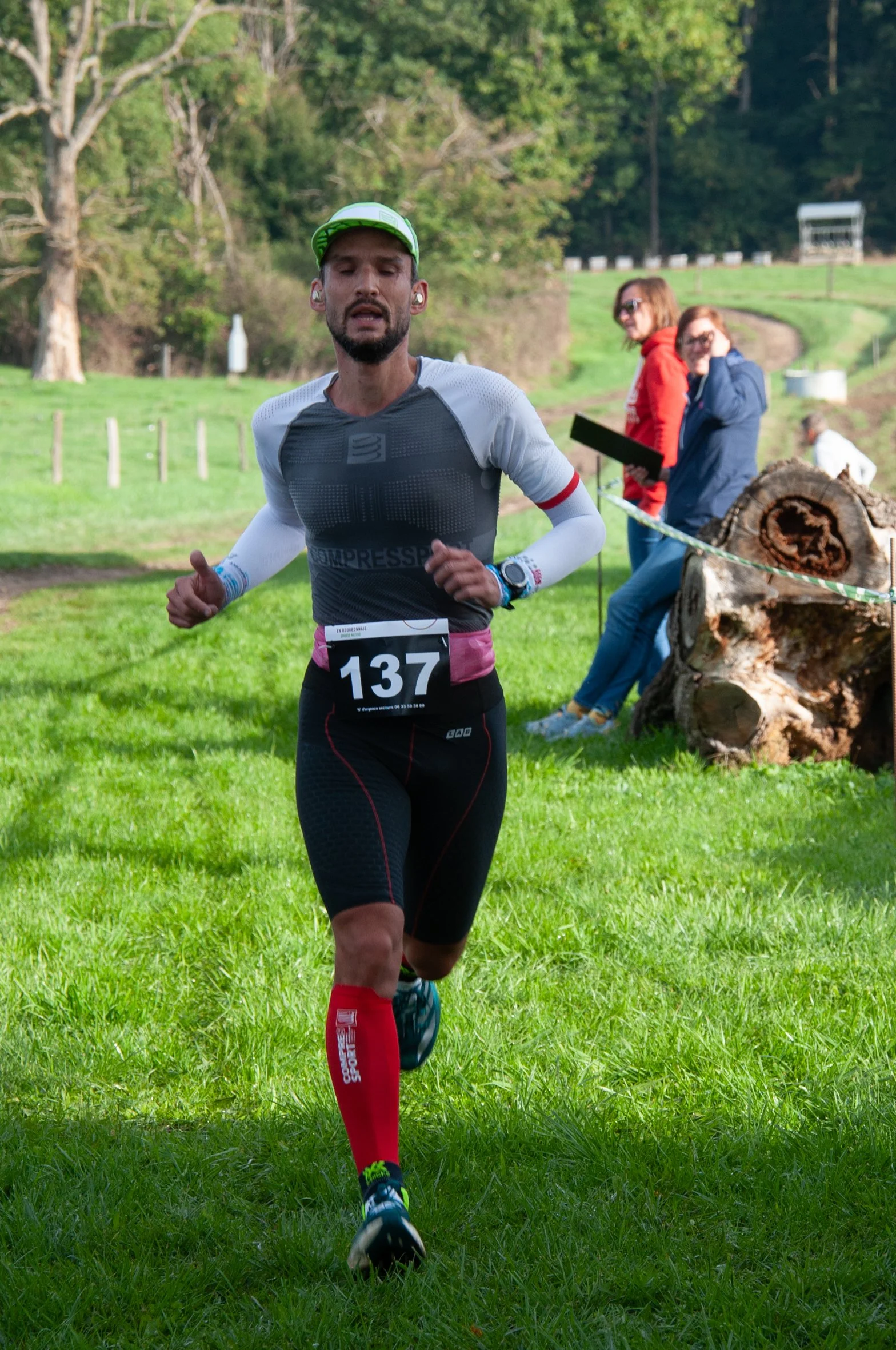 Un homme court lors d'une compétition de course à pied en plein air, portant un maillot de sport noir et blanc, un numéro 137, avec un casque de course, sur un terrain en herbe, en arrière-plan des spectateurs assis près d’un tronc d'arbre.
