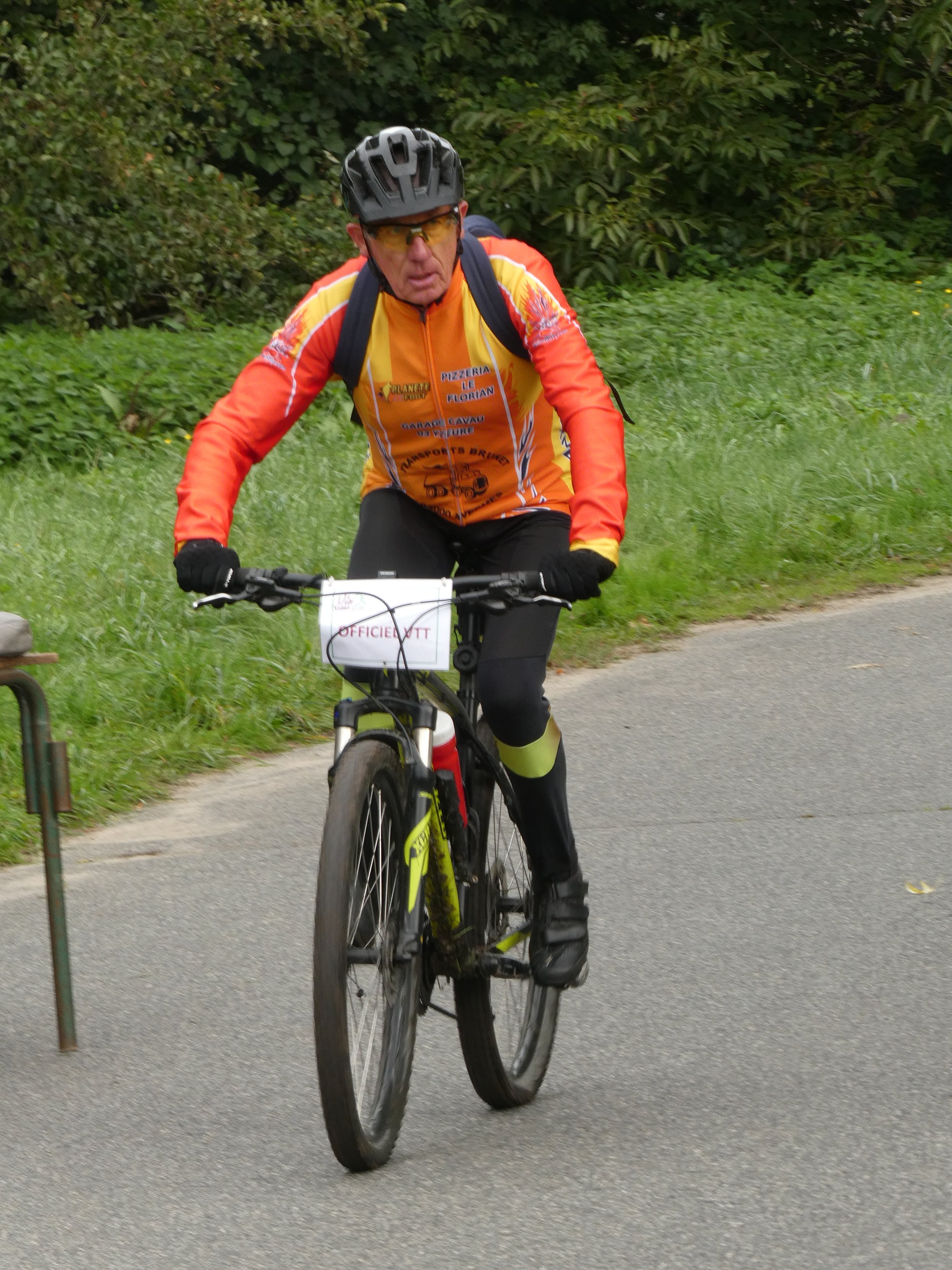 Un homme en haut de vélo, portant un casque noir, des lunettes de soleil jaunes, une veste orange et noire, et des gants noirs, en train de faire du vélo sur un chemin vertical avec de la végétation verte sur le côté.