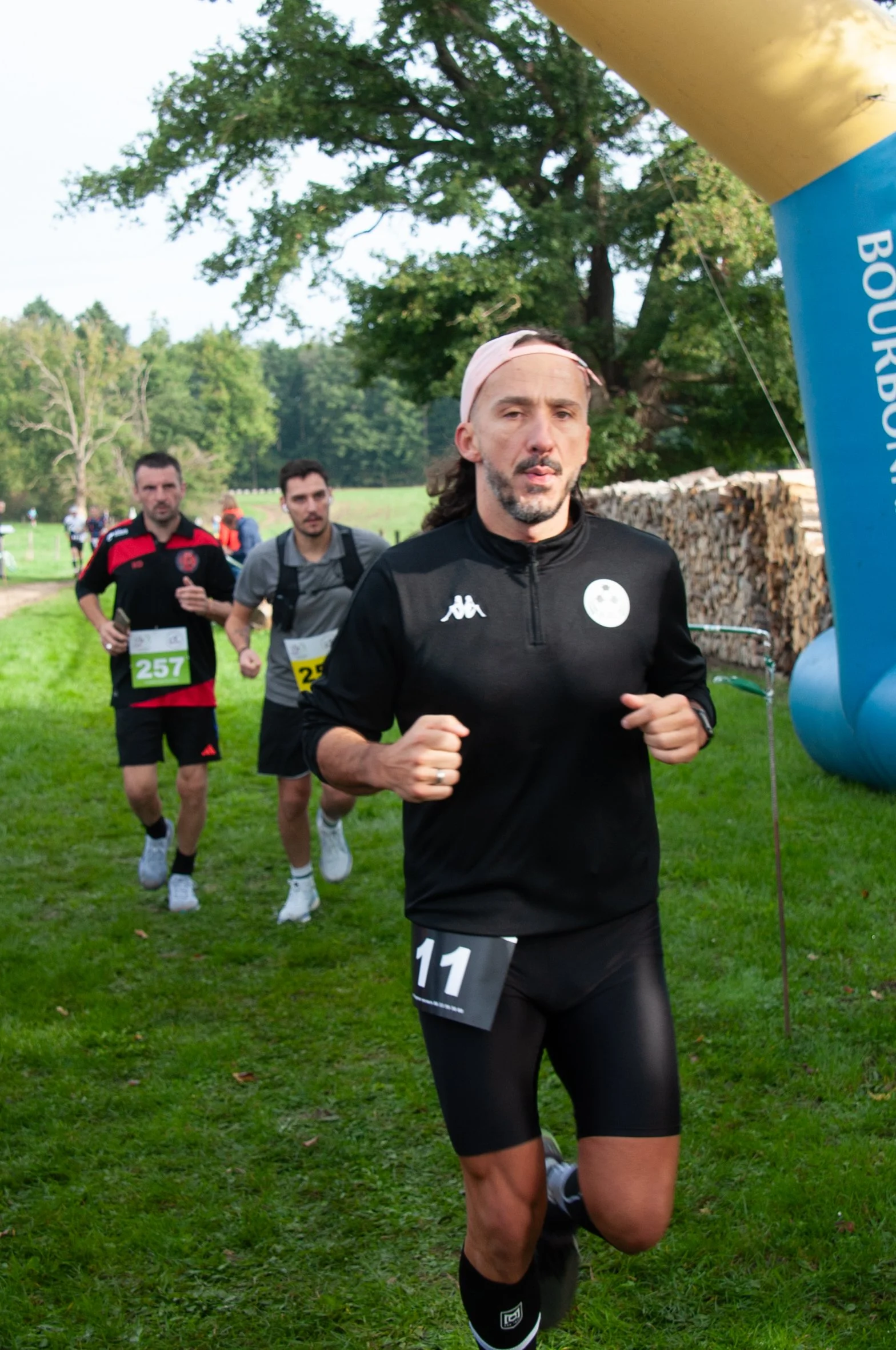Groupe de coureurs participant à une course en plein air, avec un homme en premier plan portant un maillot noir avec le numéro 11, courant sur une pelouse verte.